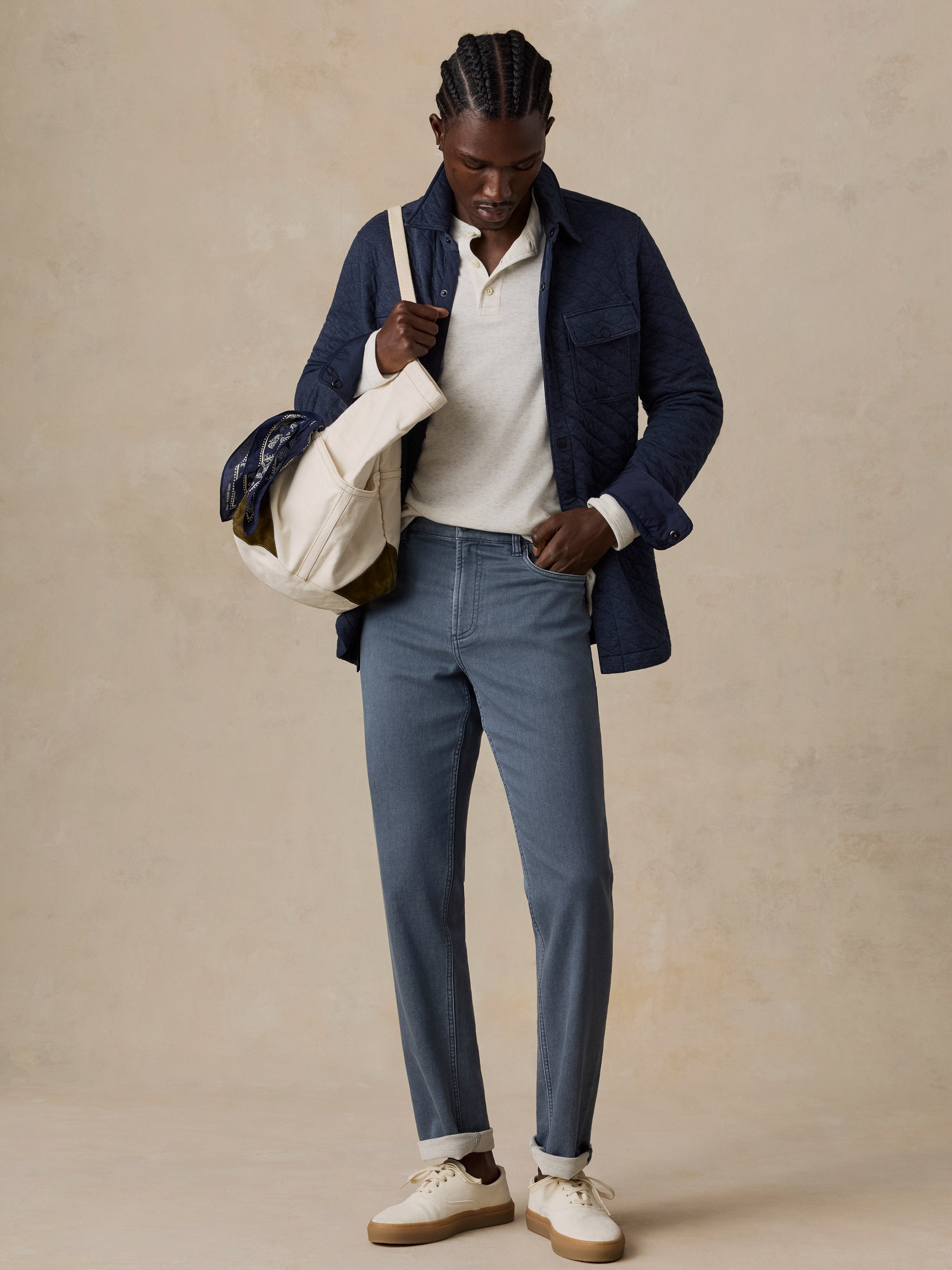 Man in navy blue quilted jacket, white polo, blue jeans, white sneakers, and cream canvas duffel bag.