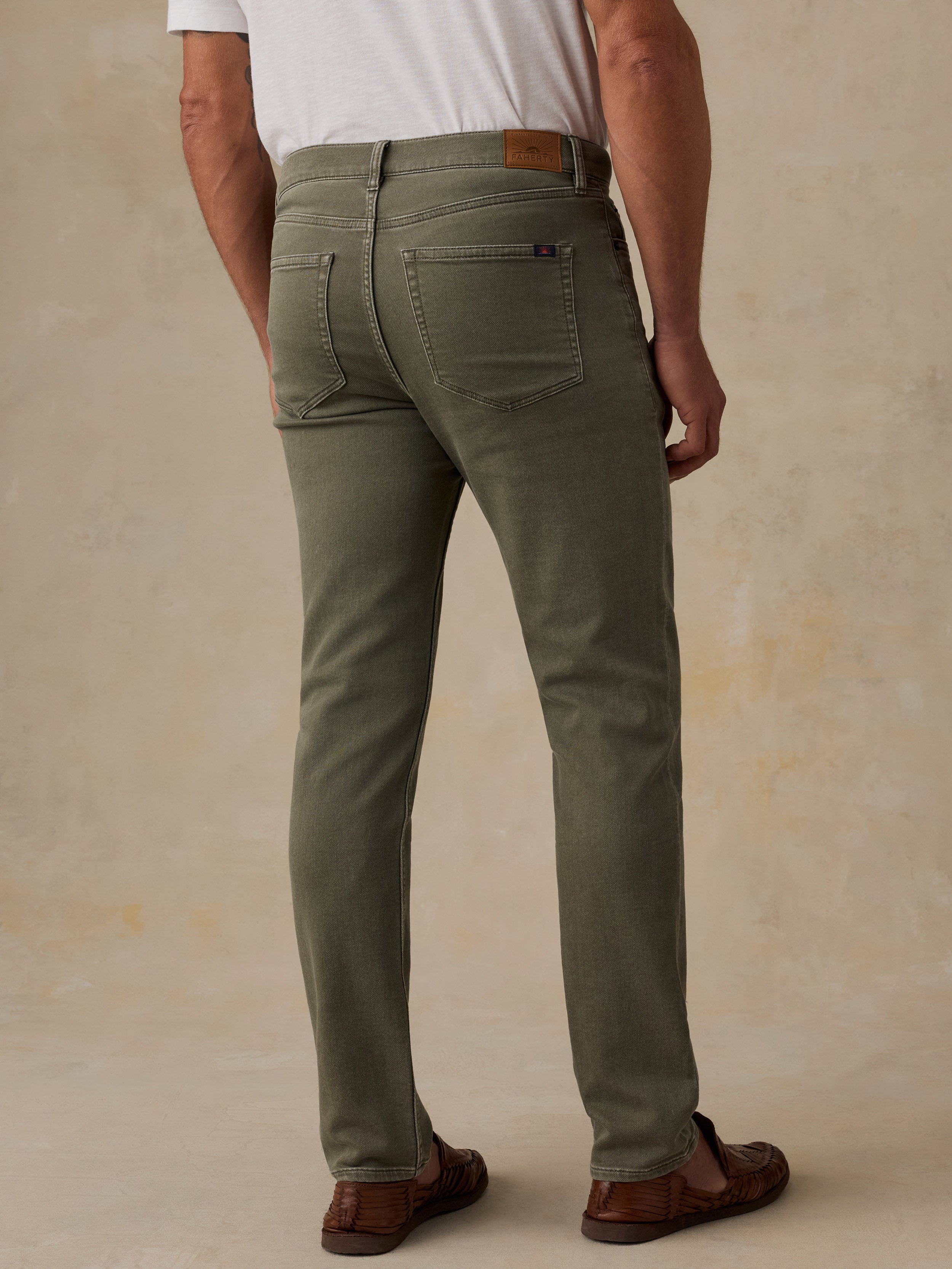 A man viewed from the back wearing olive green slim-fit jeans, a white t-shirt, and brown leather loafers, standing against a textured beige studio backdrop.