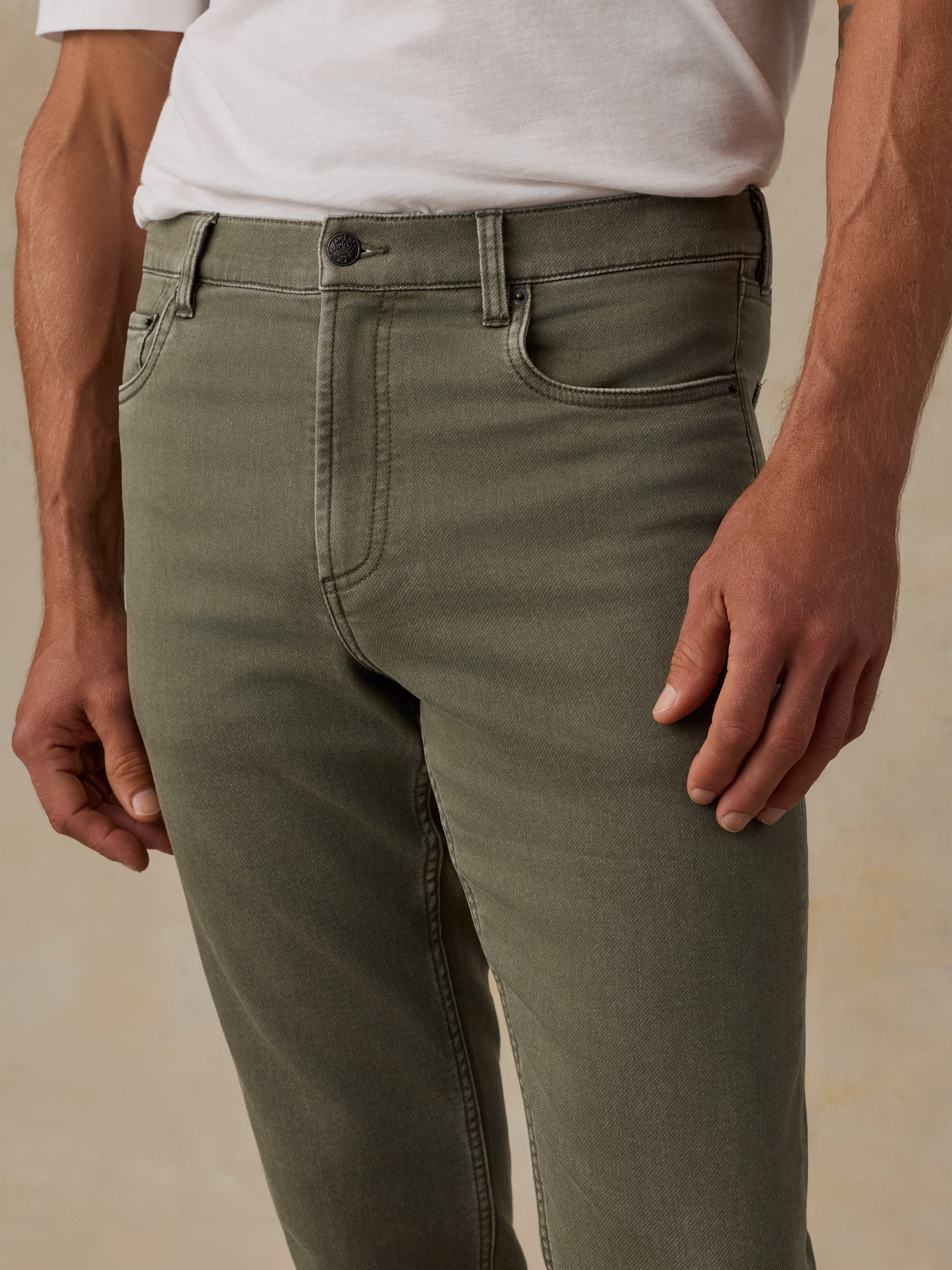 A close-up of a man wearing olive green slim-fit jeans with a white t-shirt tucked in, showing the front waistband, belt loops, button, and front pockets.