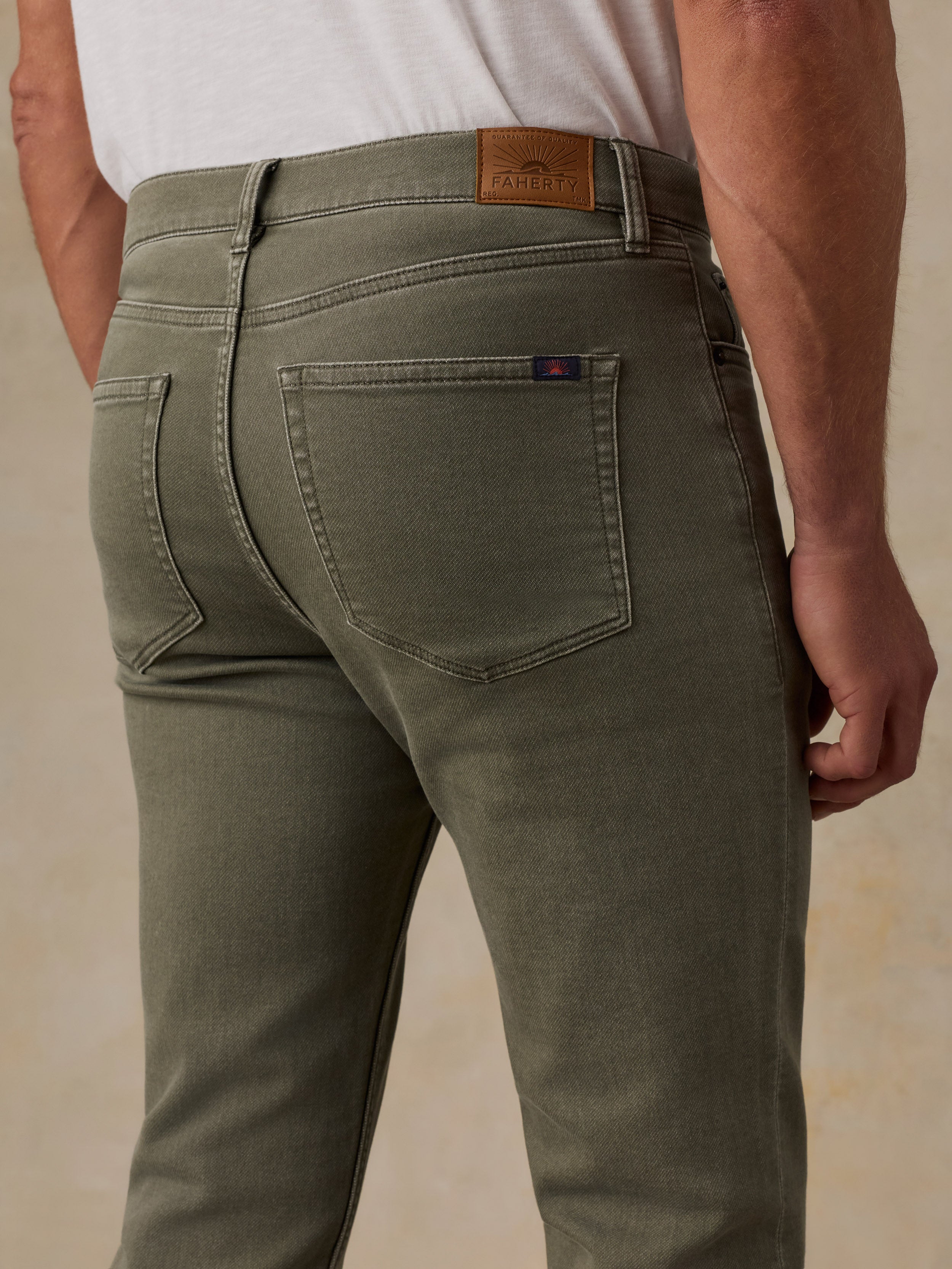 Close-up back view of a man wearing olive green jeans with a white t-shirt, showing the back pocket, waistband, and leather brand patch.
