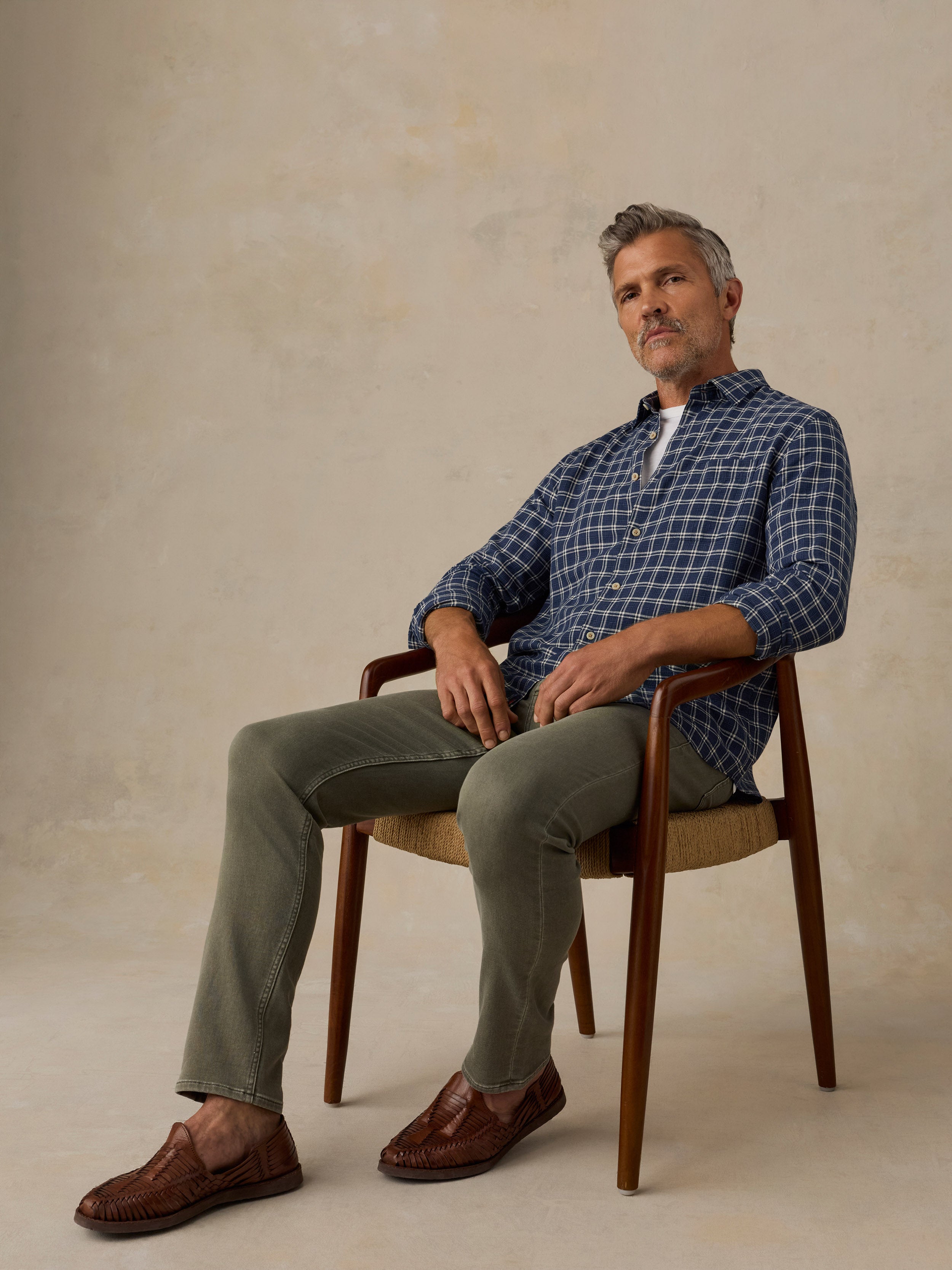 A man sits in a wooden armchair wearing a blue and white checkered long-sleeve button-up shirt, olive green trousers, and brown leather loafers against a textured beige studio backdrop.