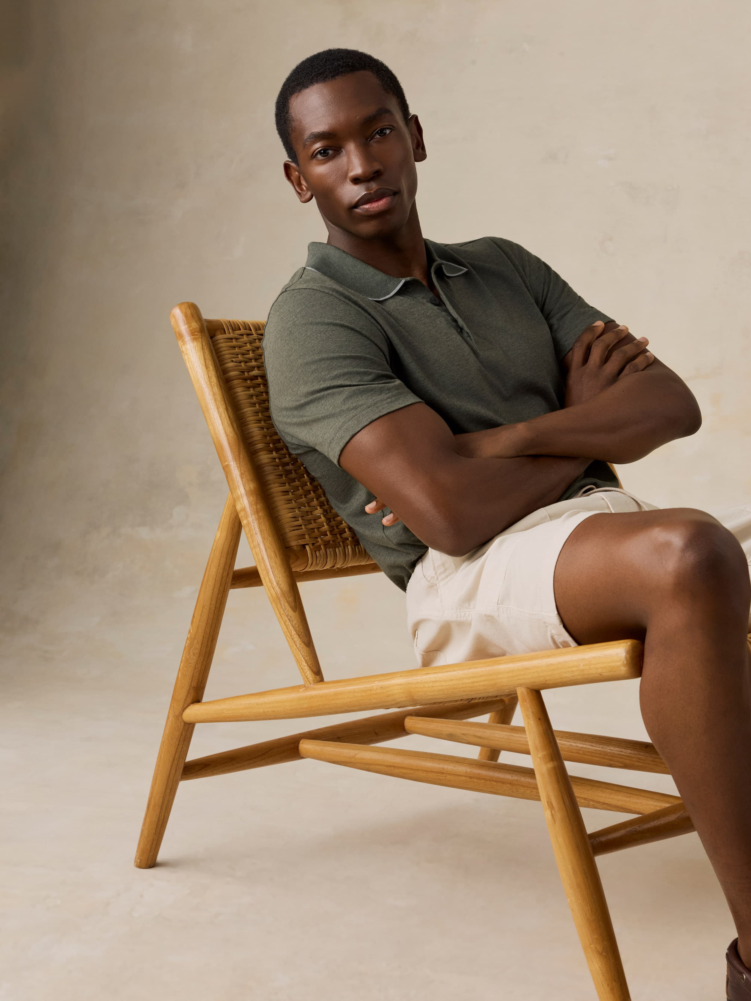 Man sitting on a wooden chair wearing a green polo shirt and beige shorts against a beige background