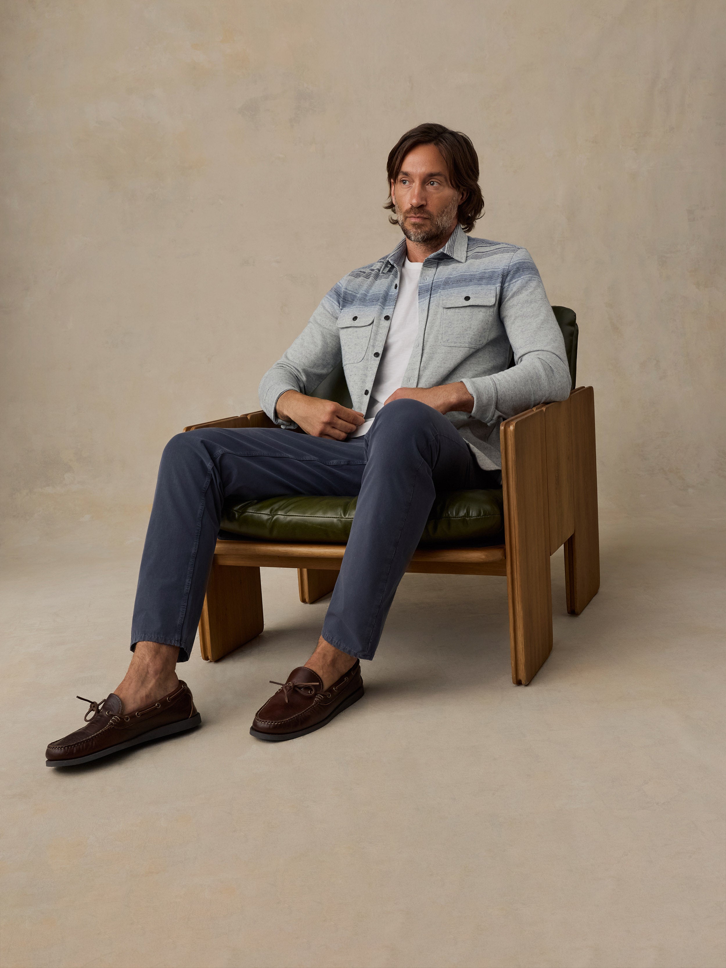 A man sits in a wooden armchair wearing a light blue denim jacket over a white shirt, navy trousers, and dark brown loafers, against a textured beige studio backdrop.