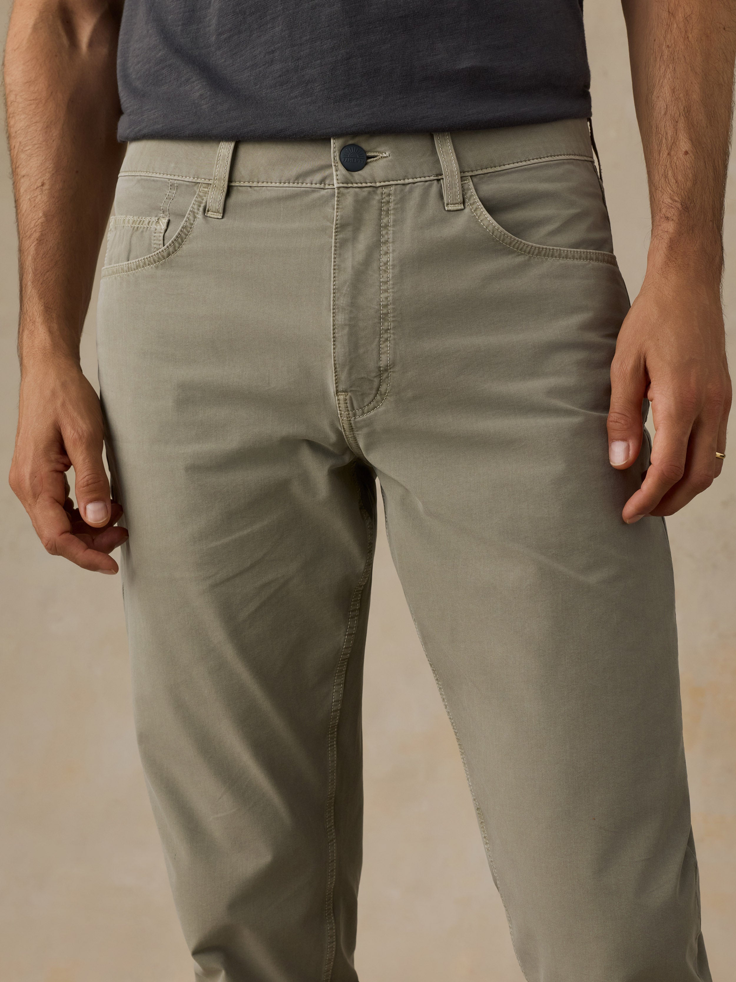 A close-up of a man wearing light olive green chino pants, showing the front waistband, belt loops, front pockets, and upper legs against a plain beige studio background.