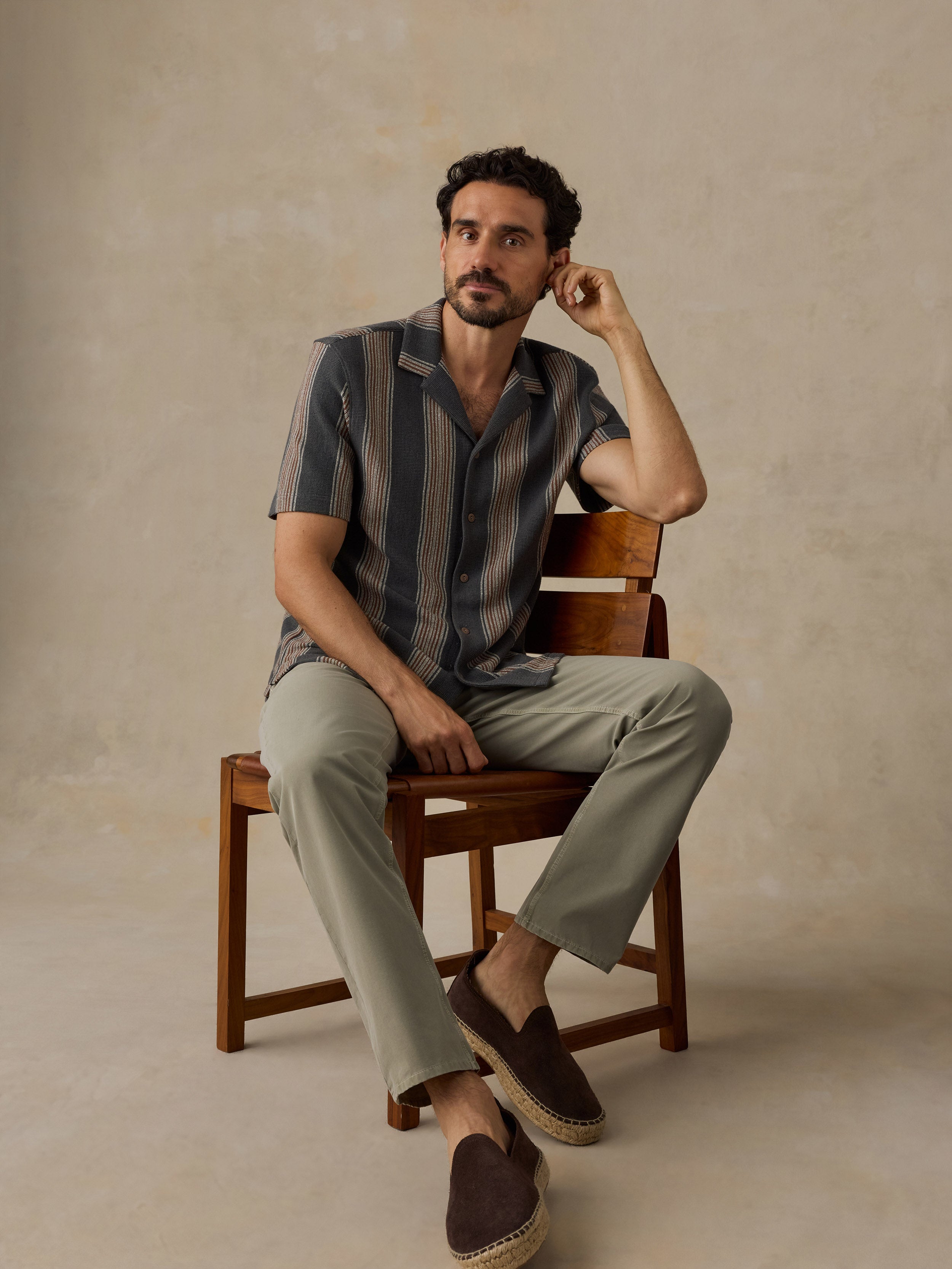 A man sits on a wooden chair wearing a short-sleeve button-up shirt with vertical stripes in dark blue and beige, light olive-green trousers, and dark brown espadrille slip-on shoes against a textured beige studio backdrop.