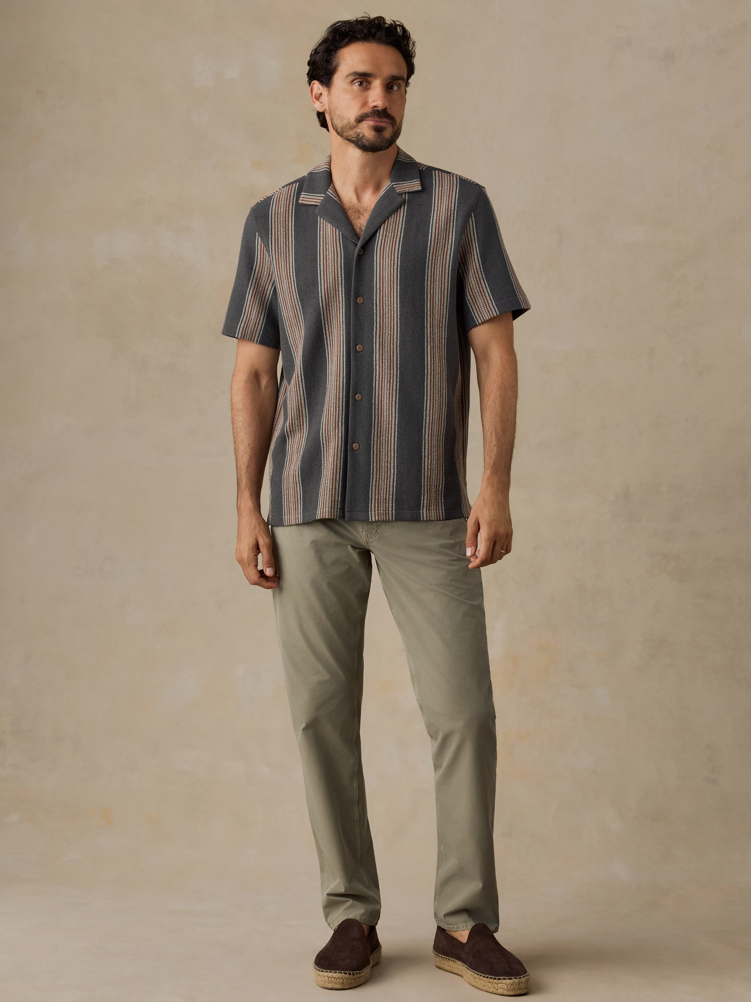 A man modeling a short-sleeve button-up shirt with vertical stripes in slate blue, gray, and beige, paired with olive green trousers and brown slip-on shoes, against a textured beige studio backdrop.