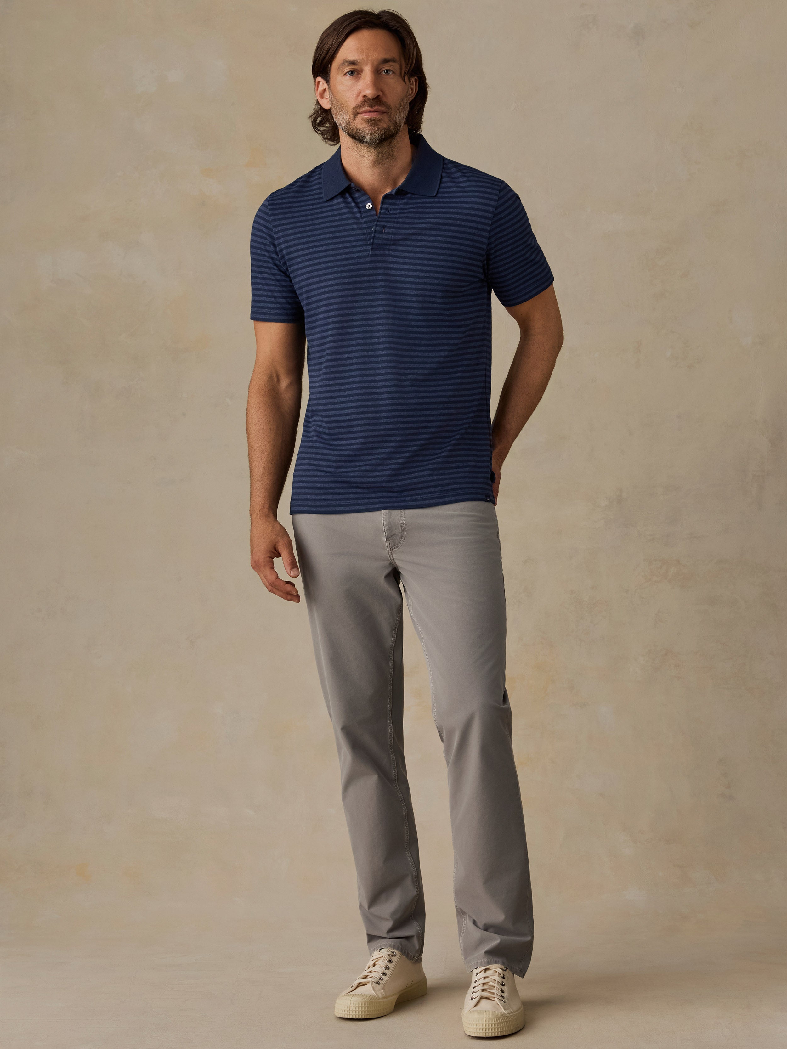 A man wearing a navy blue short-sleeve polo shirt with thin horizontal stripes, paired with light gray trousers and beige sneakers, standing against a textured beige studio backdrop.