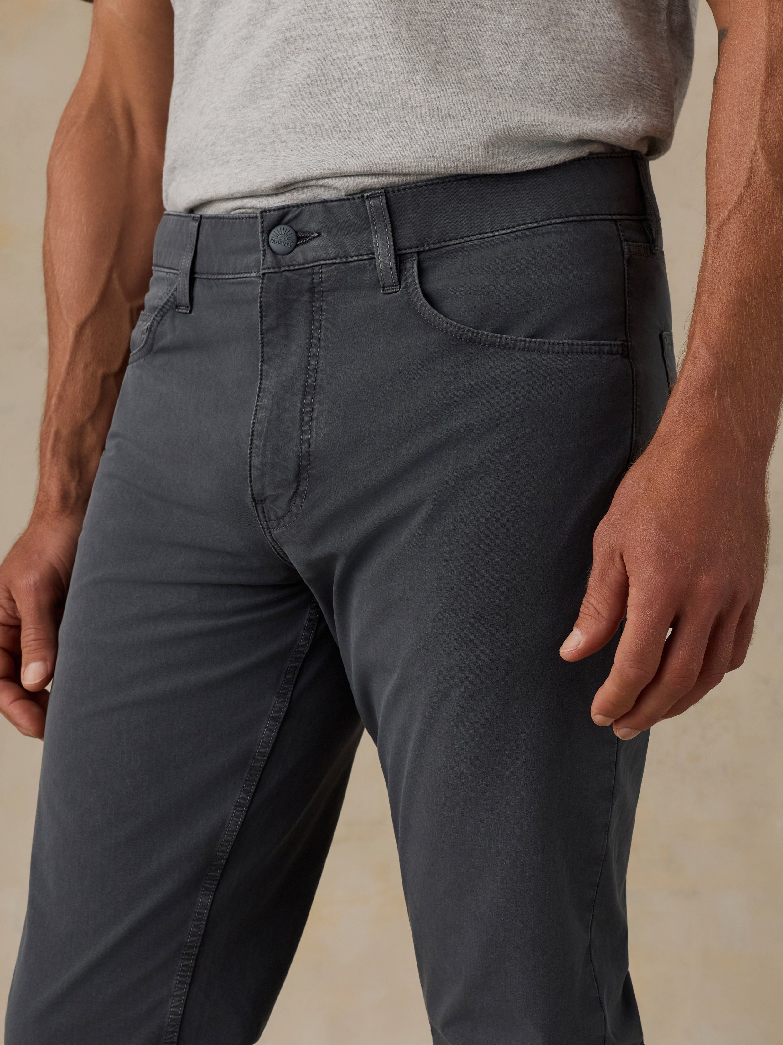 A close-up of a man wearing dark charcoal gray slim-fit pants with a heather gray t-shirt, shown from the waist down against a neutral beige background.