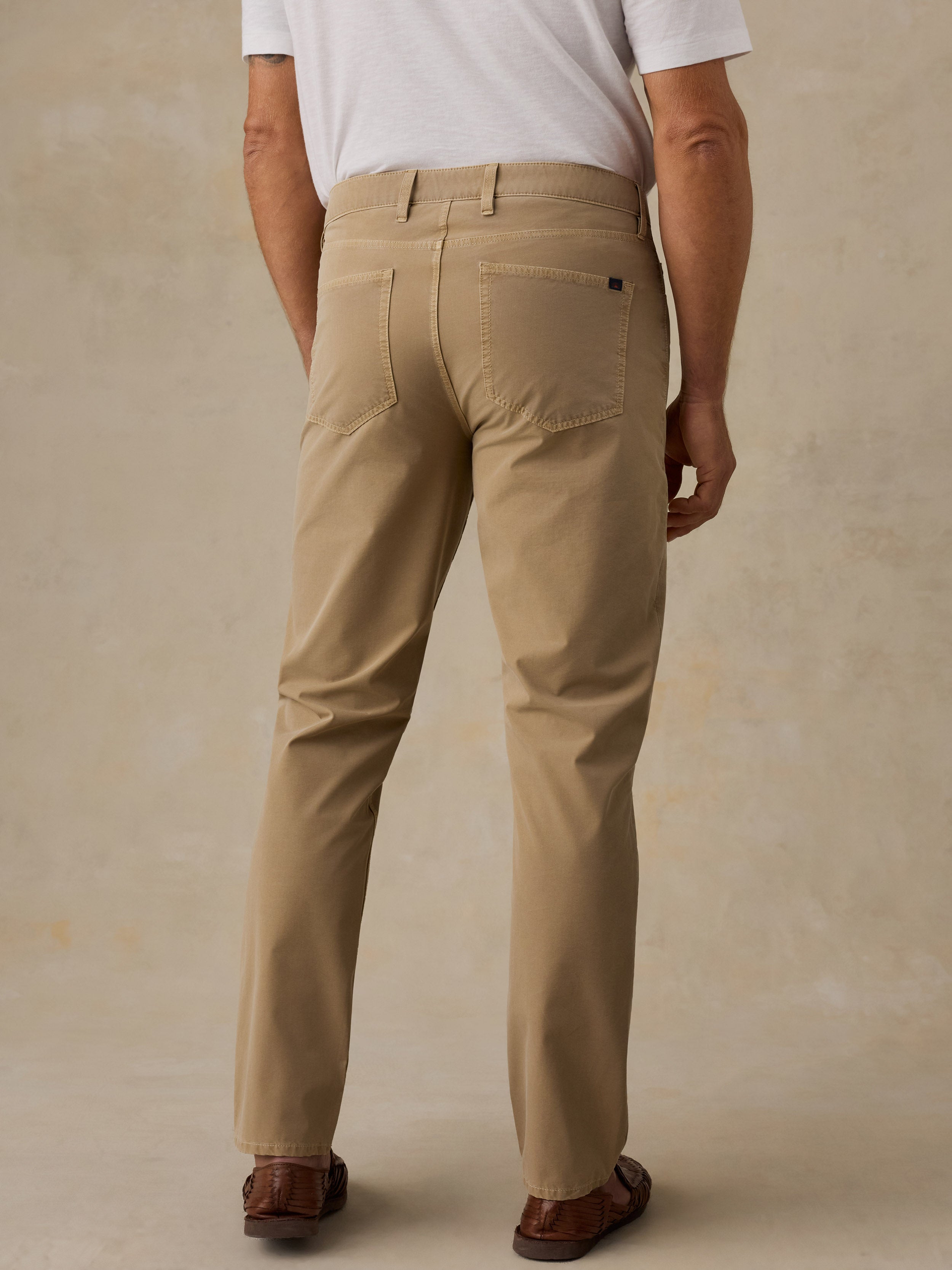 A man viewed from the back wearing straight-leg khaki trousers, a white t-shirt, and brown leather sandals, standing against a textured beige studio background.