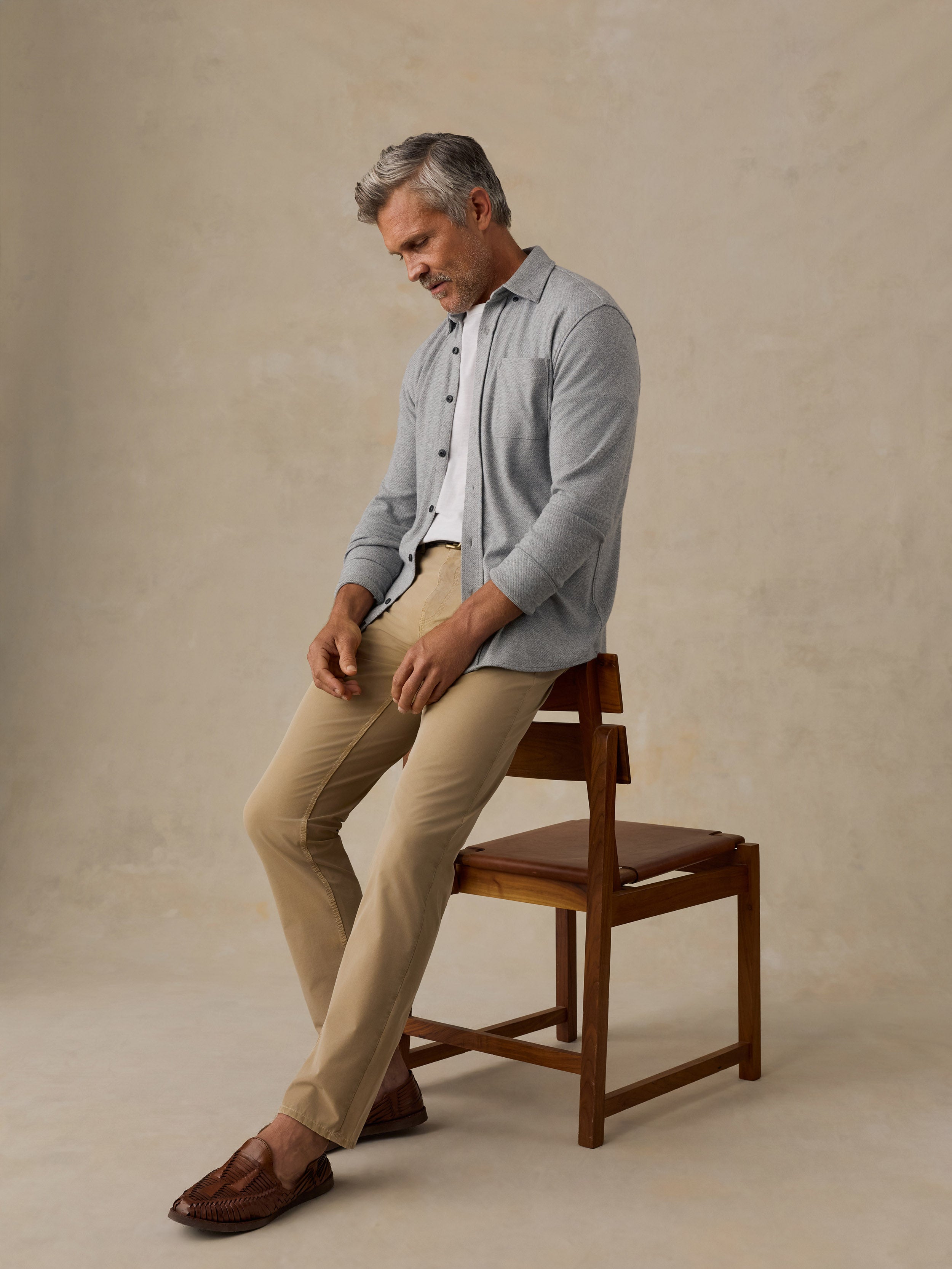 A man with gray hair sits on a wooden chair, wearing a light blue button-up shirt over a white undershirt, beige trousers, and brown loafers, against a textured beige studio backdrop.