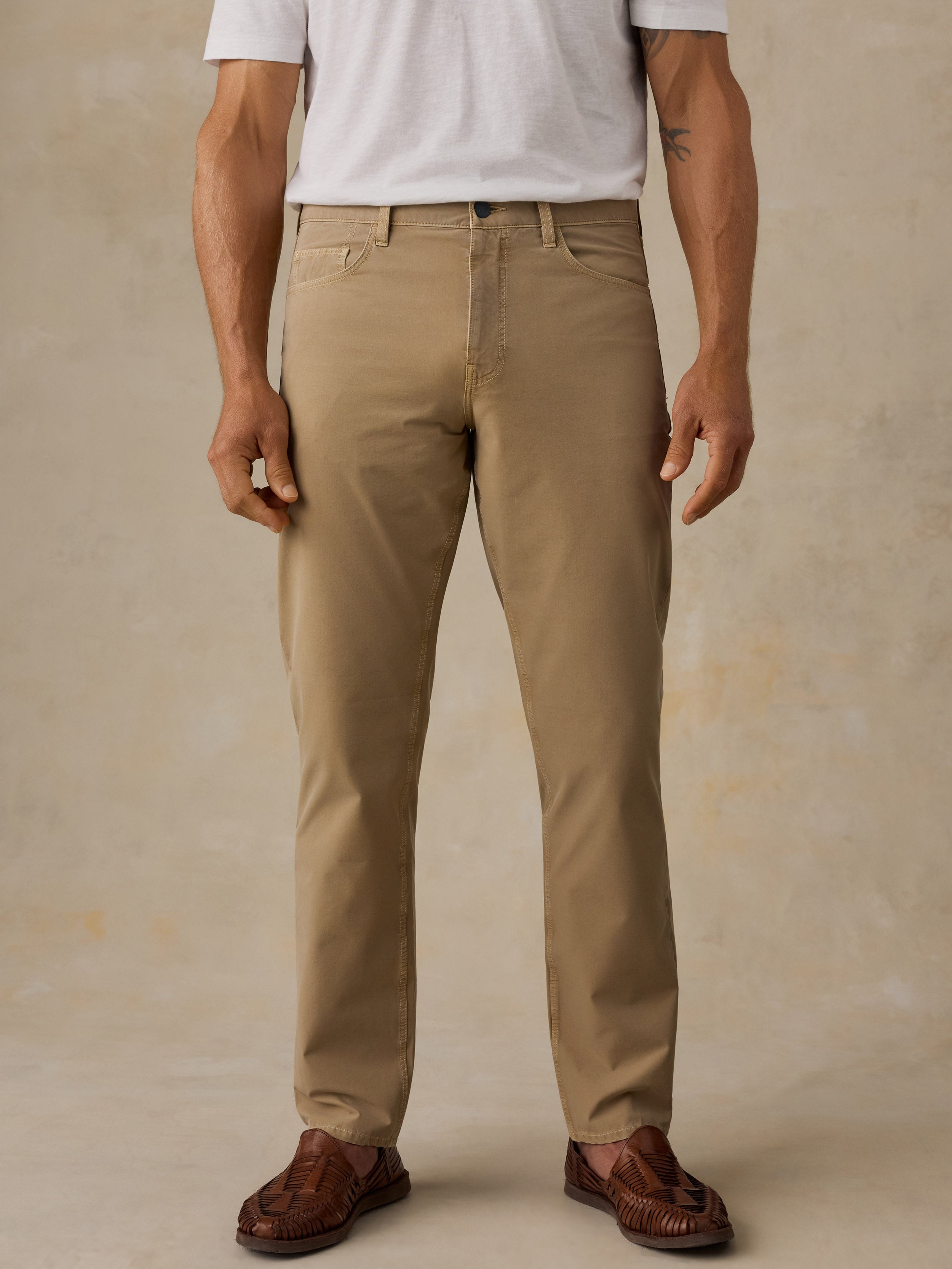 A man wearing khaki chino pants, a white t-shirt, and brown leather loafers, standing against a textured beige studio backdrop.