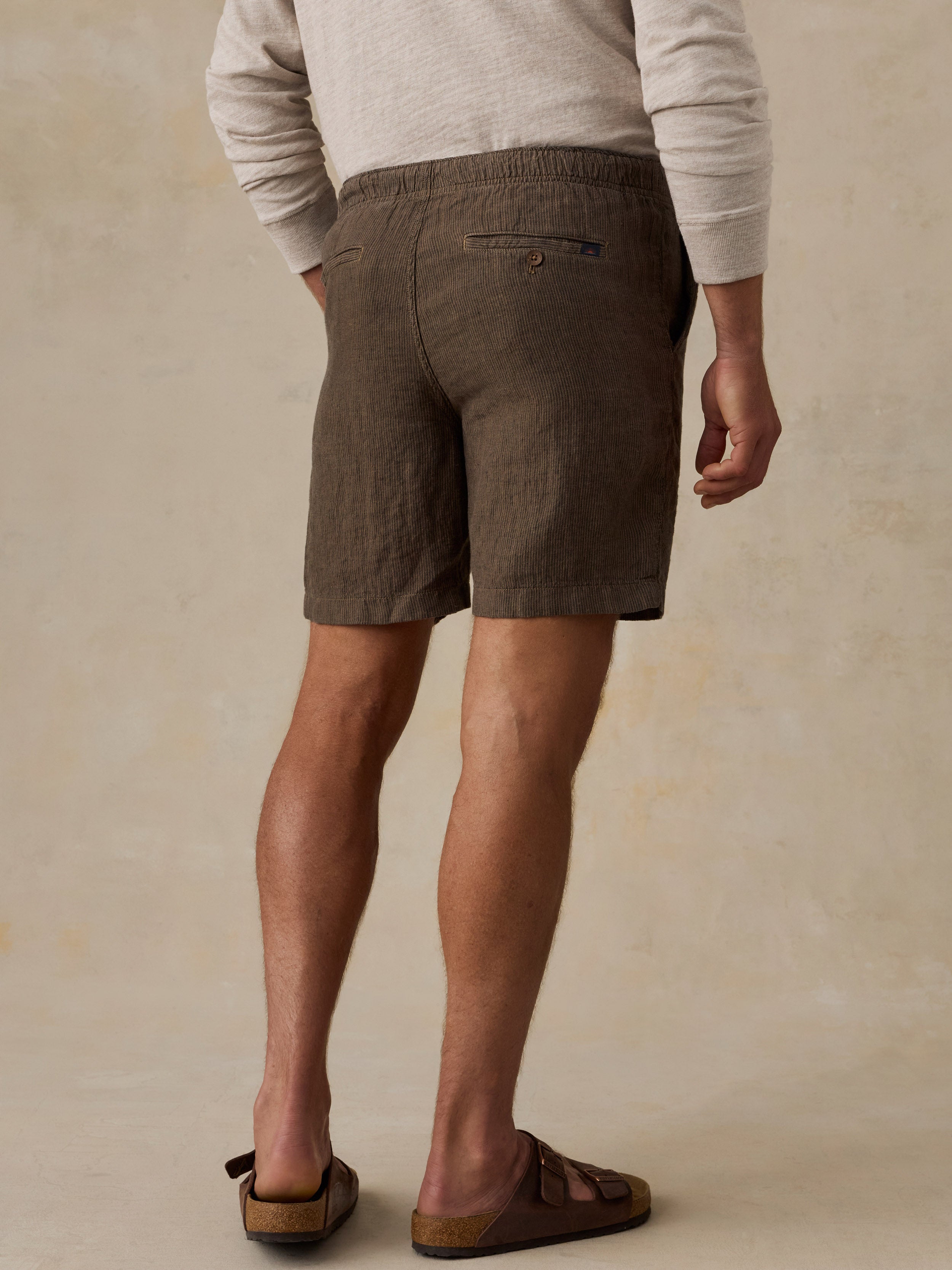 A man wearing brown linen shorts, a light beige knit sweater, and brown leather slide sandals, shown from a rear three-quarter angle against a textured beige studio backdrop.