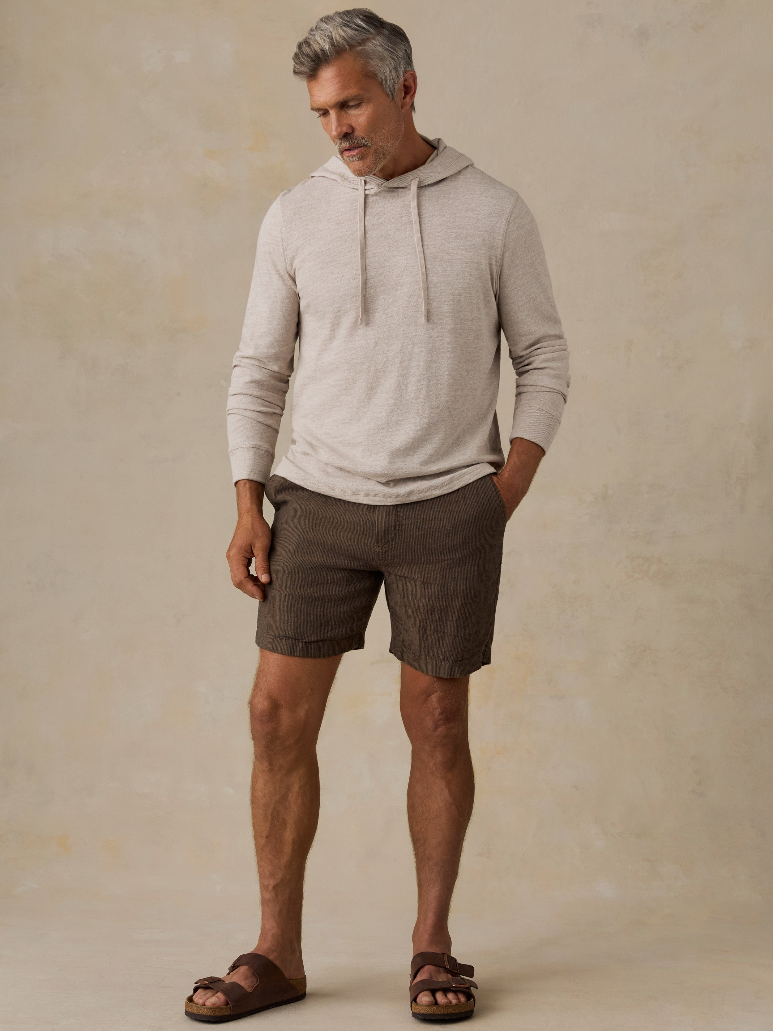 A man wearing a light beige long-sleeve hoodie and dark brown linen shorts, standing against a textured beige studio backdrop.