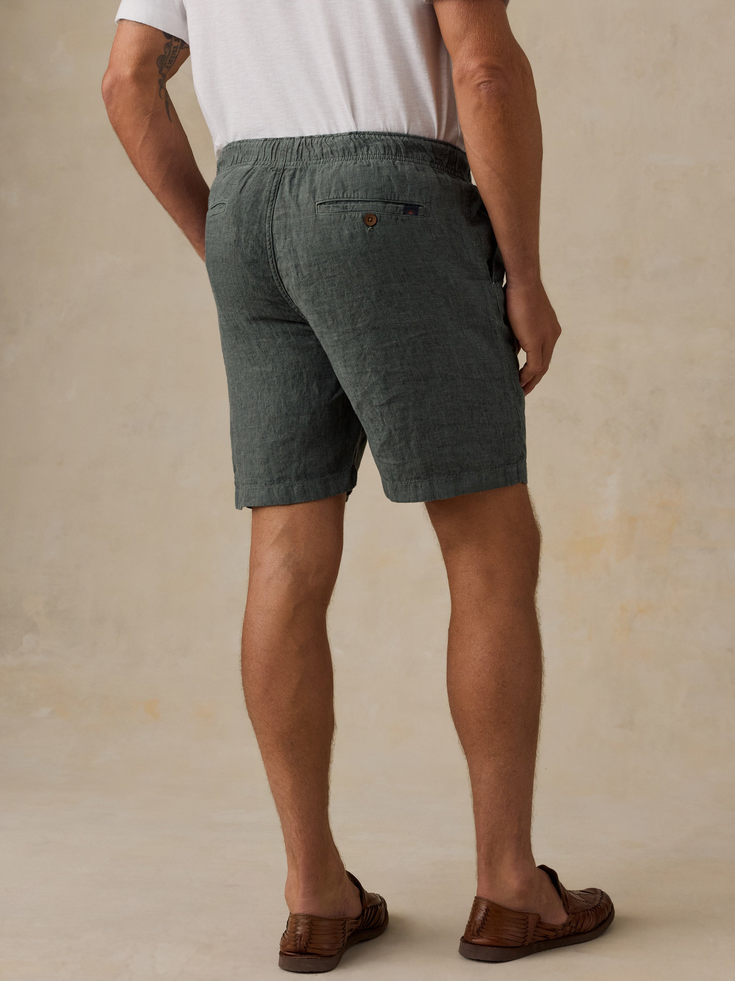 A man viewed from the back wearing dark green textured linen shorts, a white t-shirt, and brown leather loafers, set against a warm beige textured studio background.