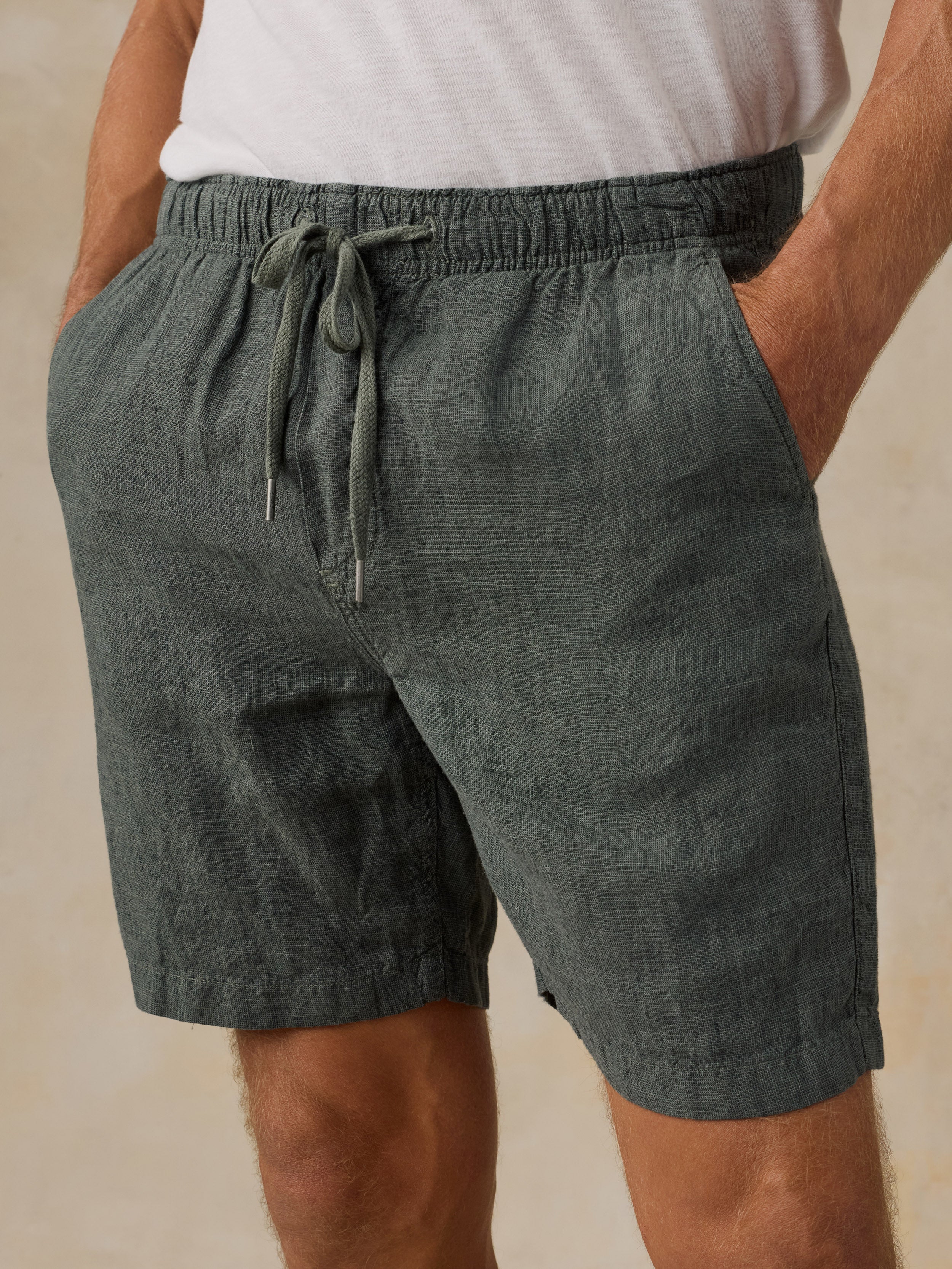 A close-up of a man wearing dark green drawstring shorts with a white t-shirt, standing against a neutral beige background.