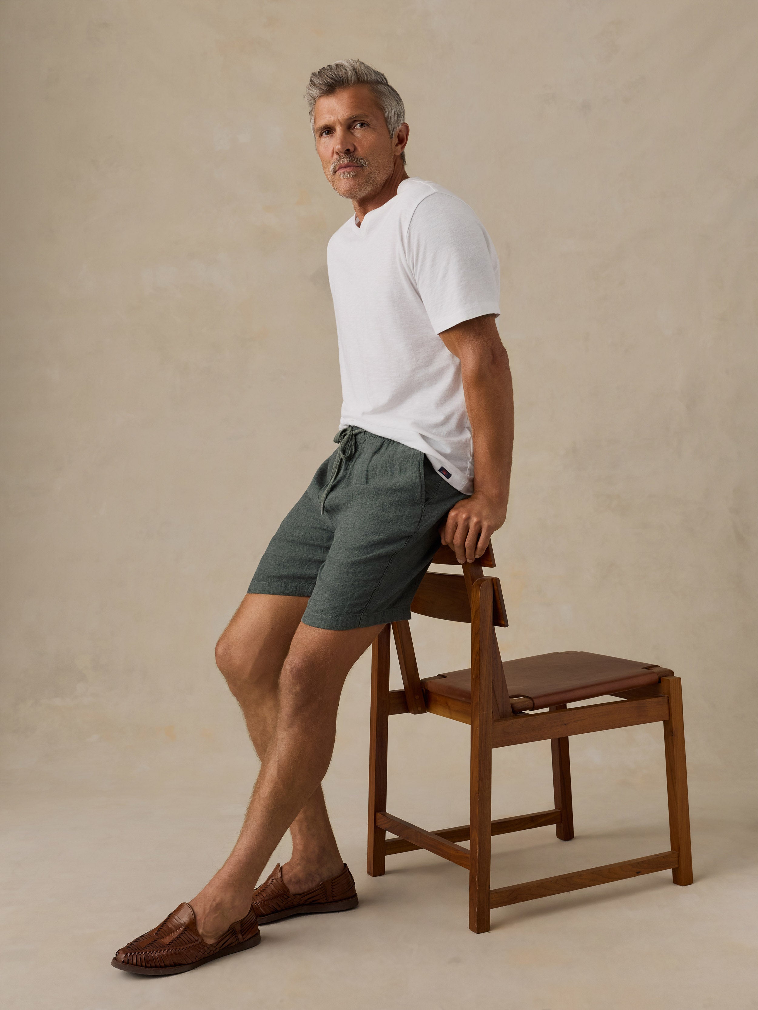 A man wearing a white short-sleeve polo shirt and dark green drawstring shorts leans against a wooden chair in a studio setting with a textured beige background.