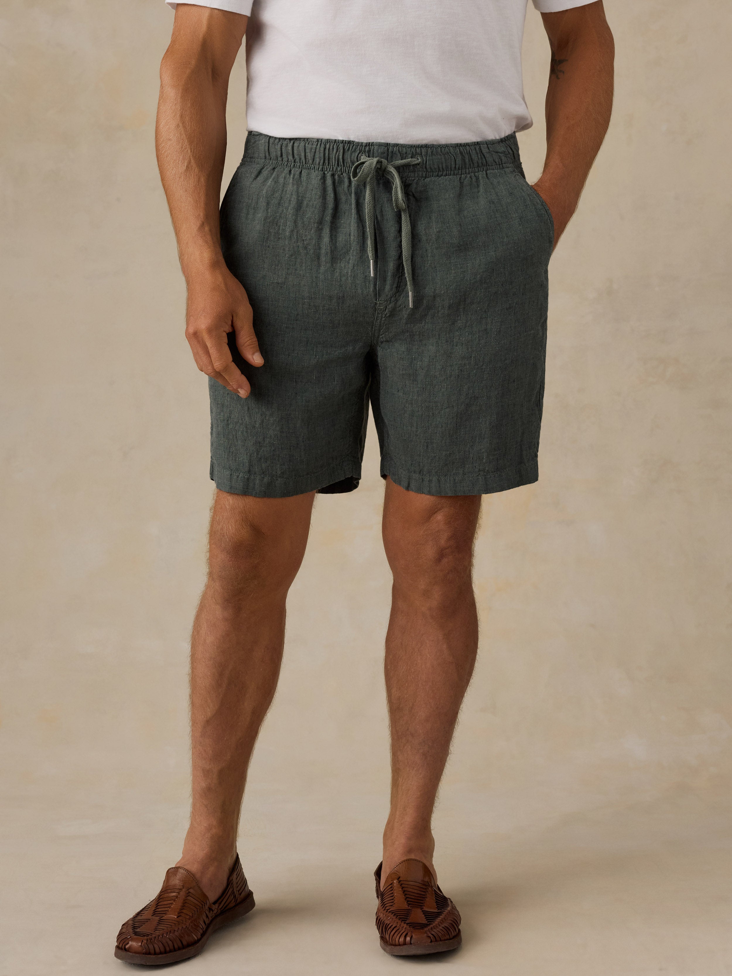 A man wearing dark green linen drawstring shorts, a white t-shirt, and brown leather loafers, standing against a textured beige studio backdrop.