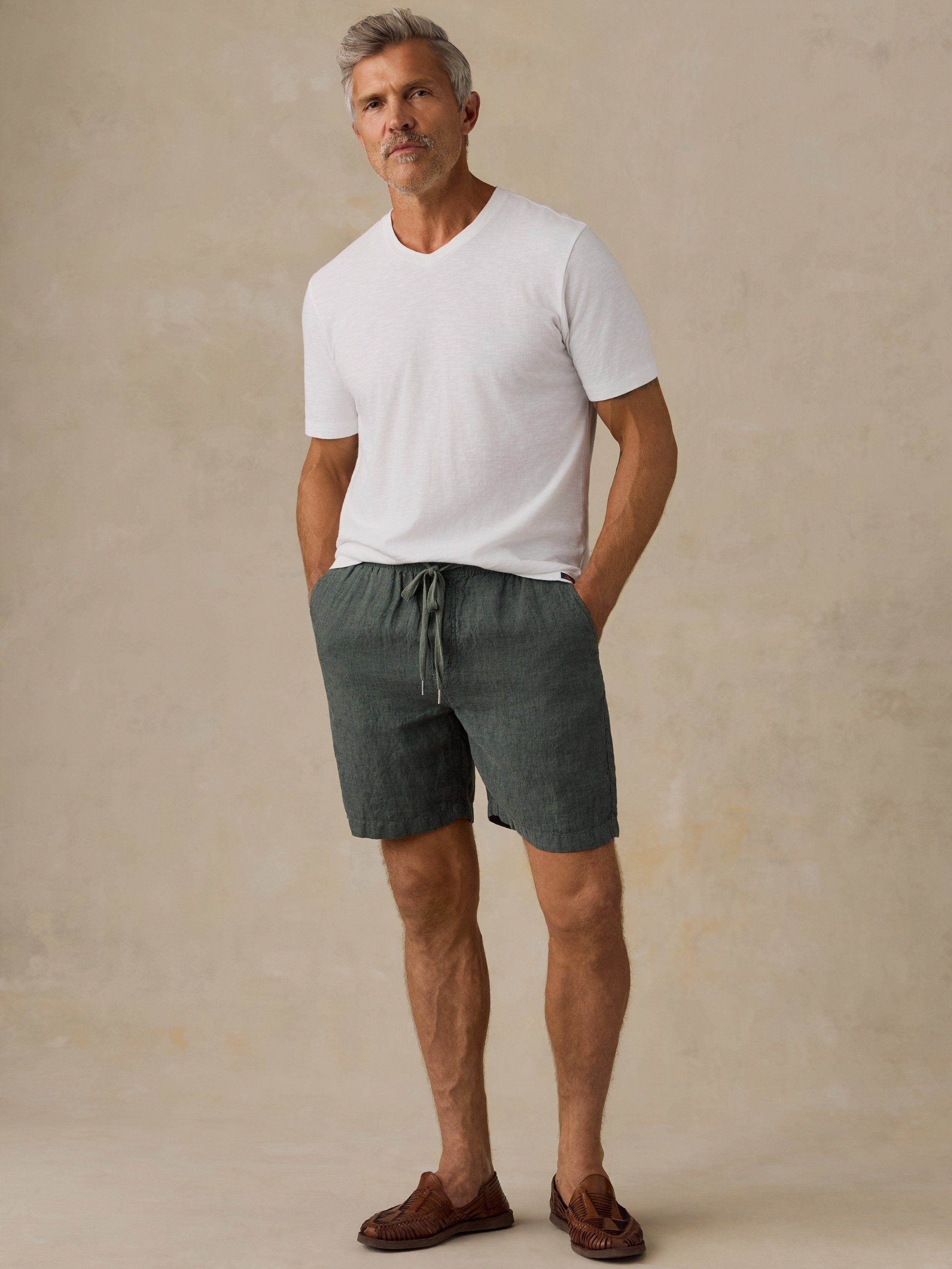 A man wearing a white short-sleeve V-neck t-shirt, dark green drawstring linen shorts, and brown leather loafers, standing against a textured beige studio backdrop.