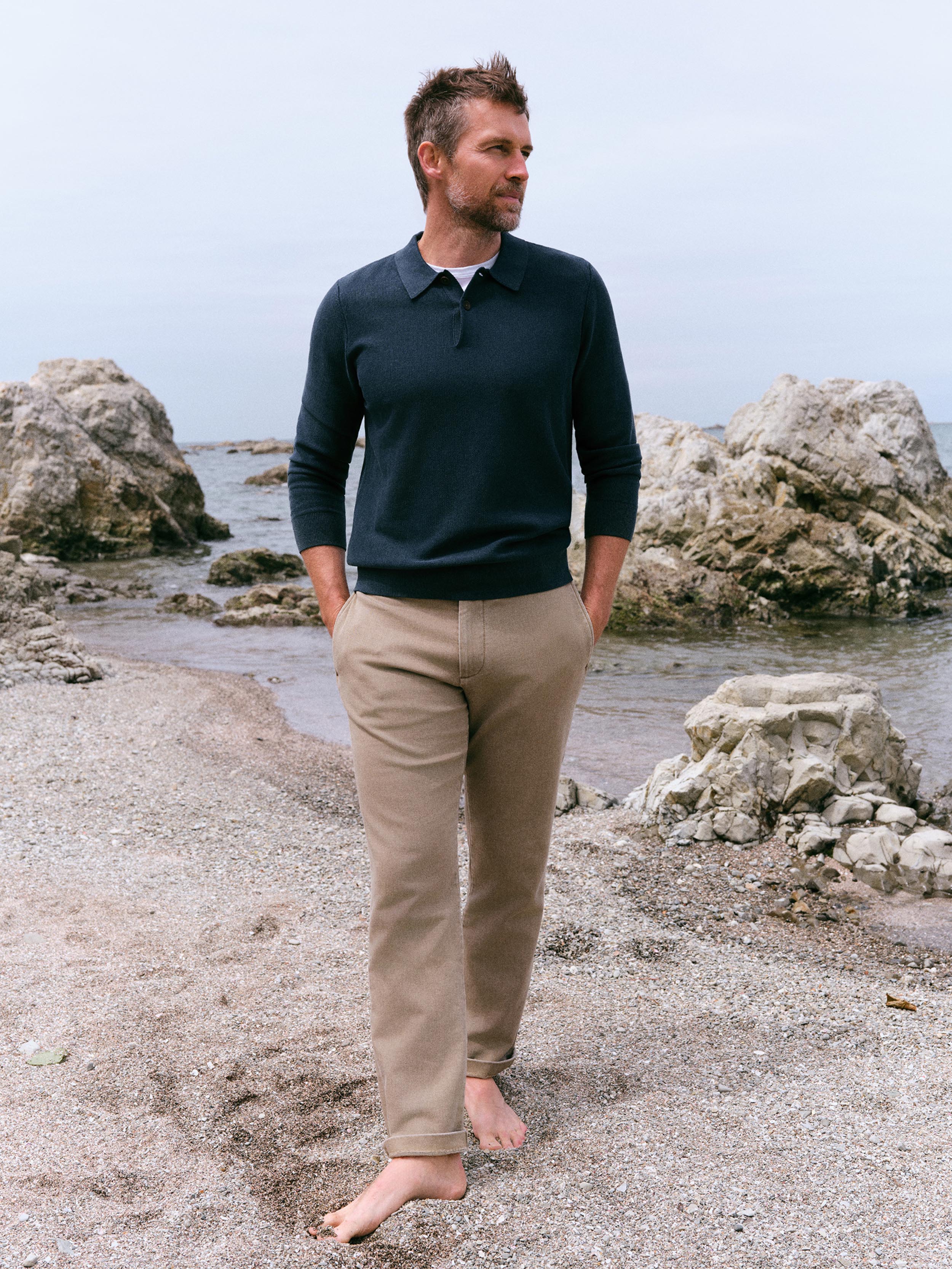 A man standing barefoot on a rocky beach shoreline, wearing a dark navy long-sleeve polo shirt and beige trousers.