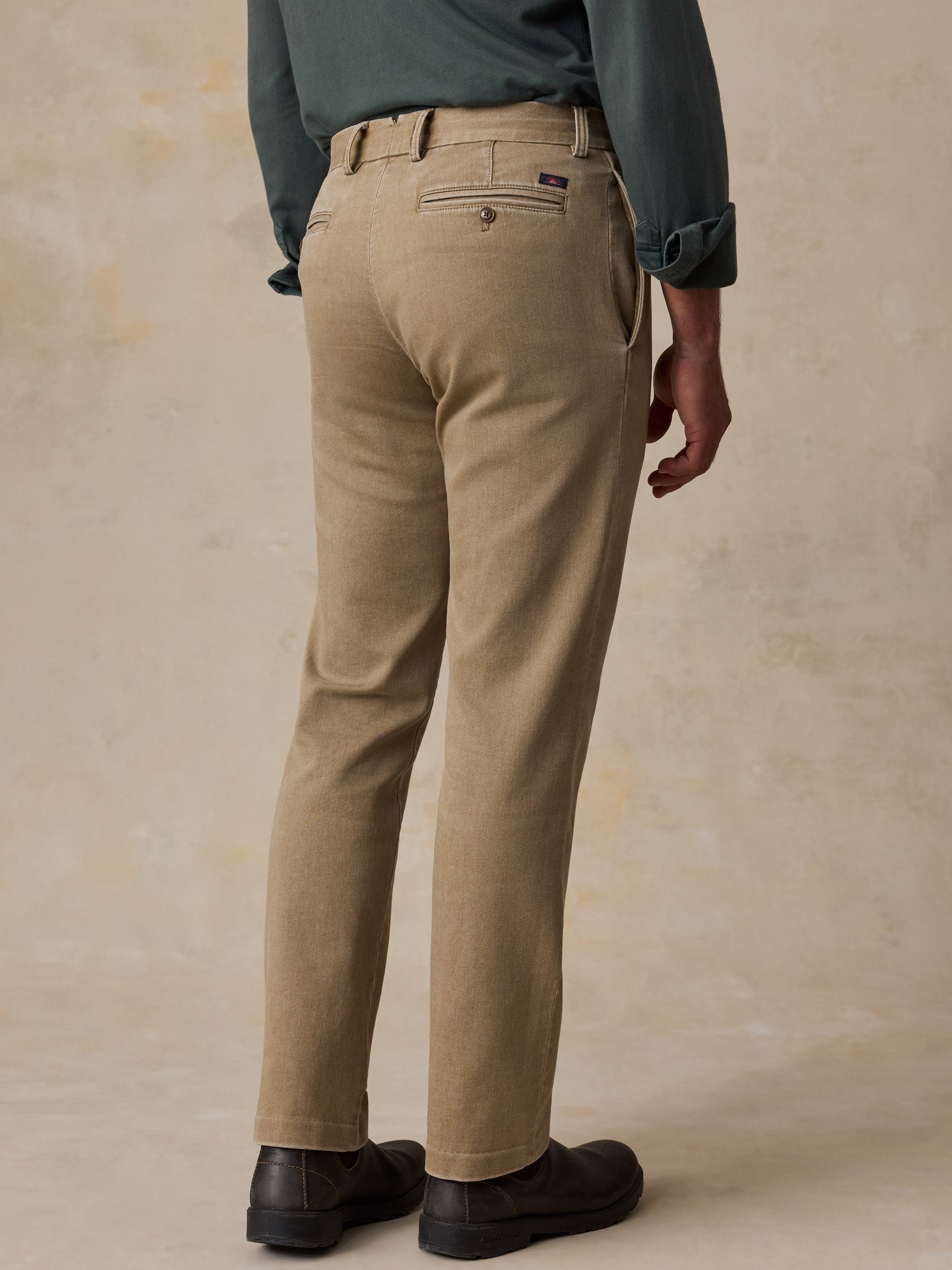 A man shown from the back wearing tan chinos with a dark green shirt and black shoes, standing against a textured beige studio backdrop.