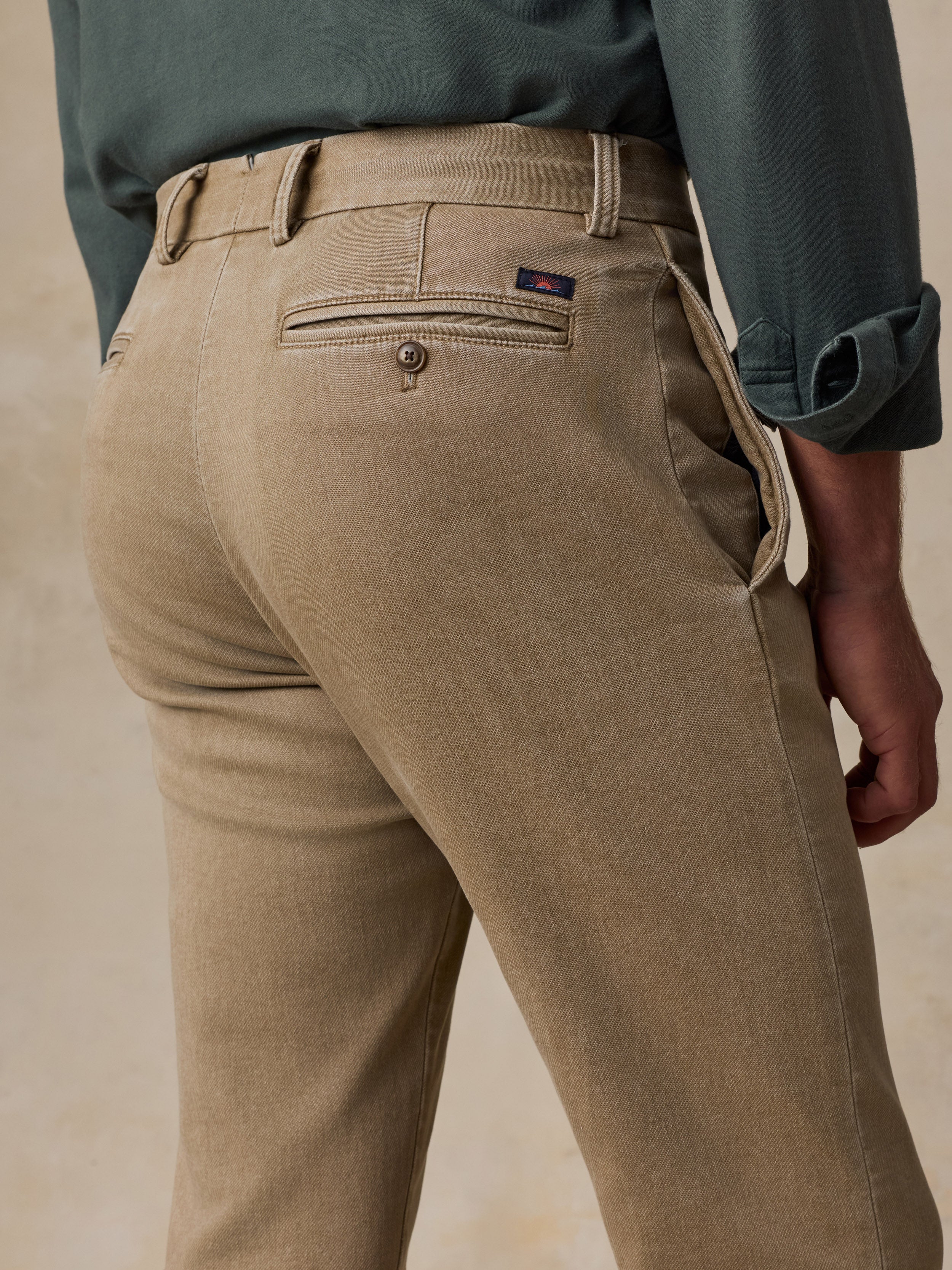 Close-up back view of a man wearing tan chinos with a belt and a dark green shirt, against a neutral beige background.