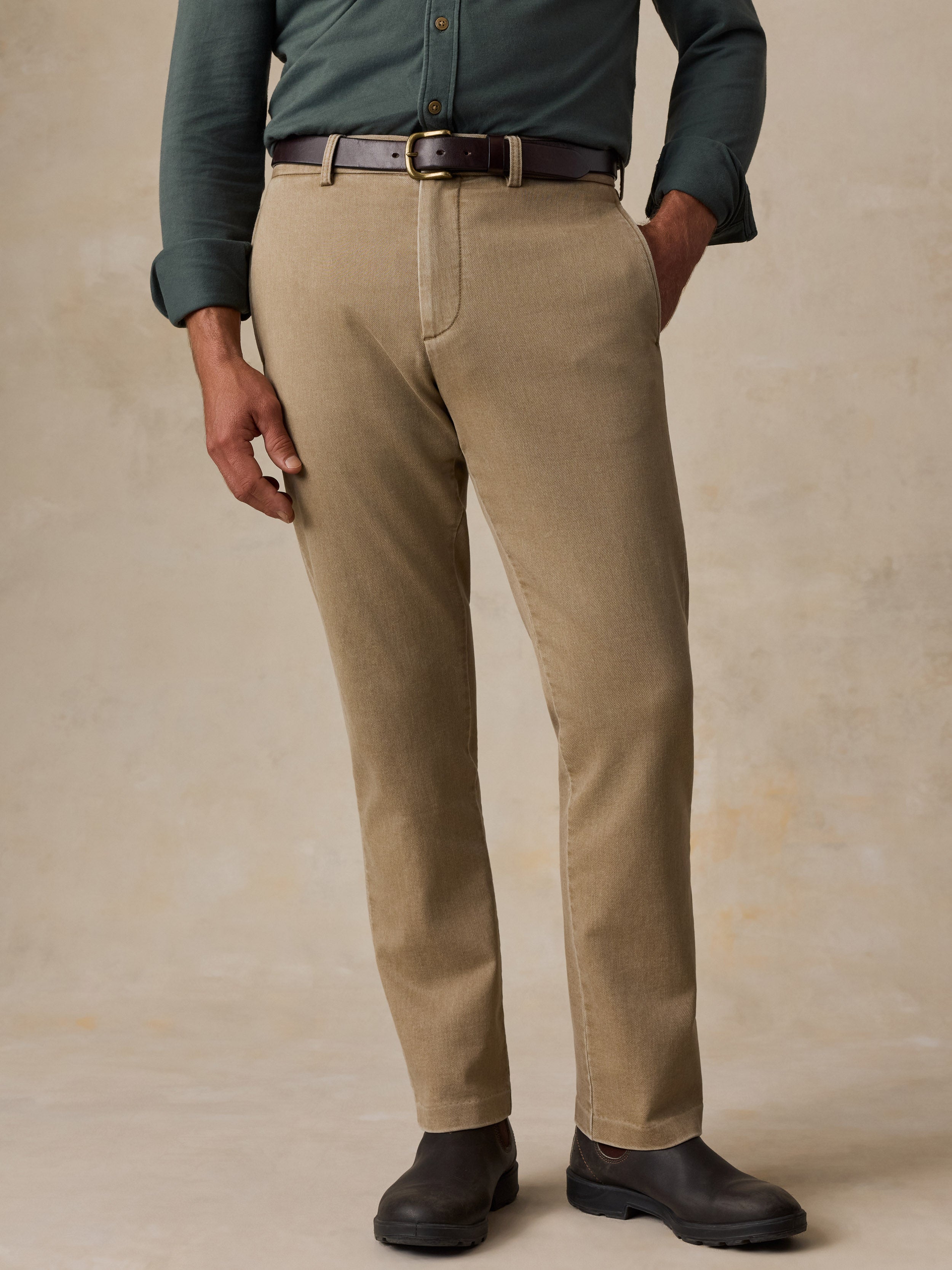 A man wearing khaki chinos with a dark brown belt, a muted green button-down shirt, and black leather boots, standing against a textured beige studio backdrop.