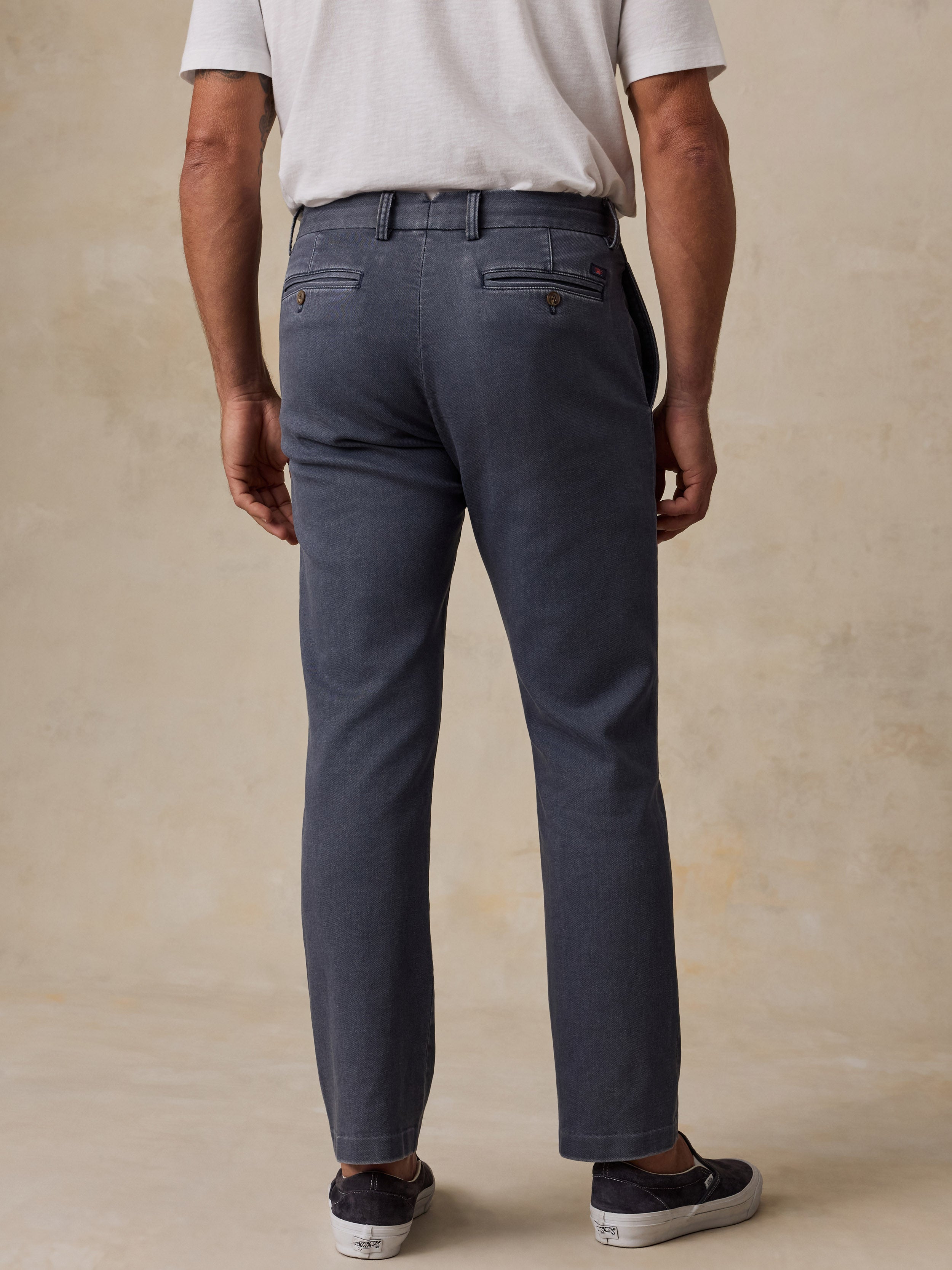 A man viewed from the back wearing slate blue chino pants, a white t-shirt, and black slip-on shoes, standing against a textured beige studio backdrop.