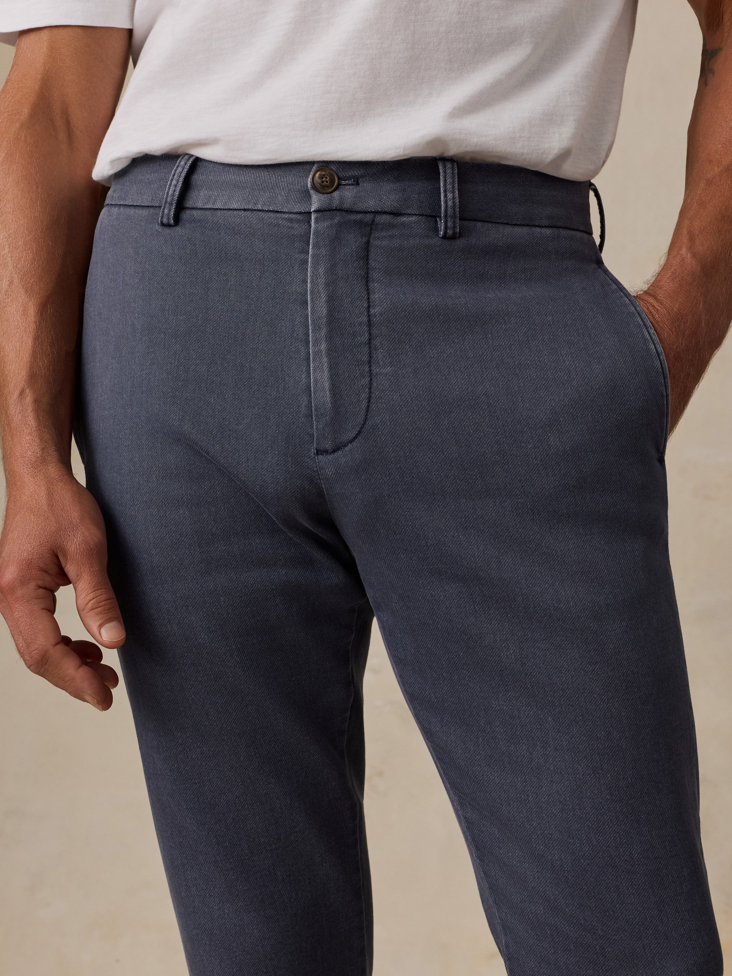 A close-up of a man wearing dark slate blue trousers with a white t-shirt, showing the front waistband, belt loops, button, and front pocket.