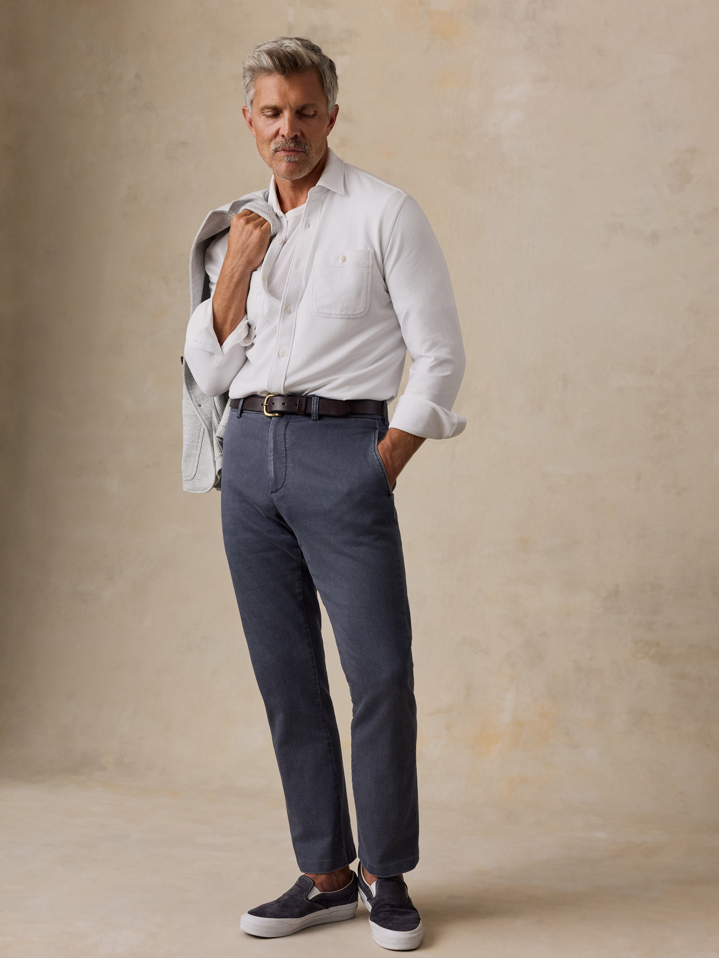 A man wearing a white long-sleeve button-down shirt, navy blue trousers, a black belt, and navy slip-on shoes, standing against a textured beige studio backdrop.