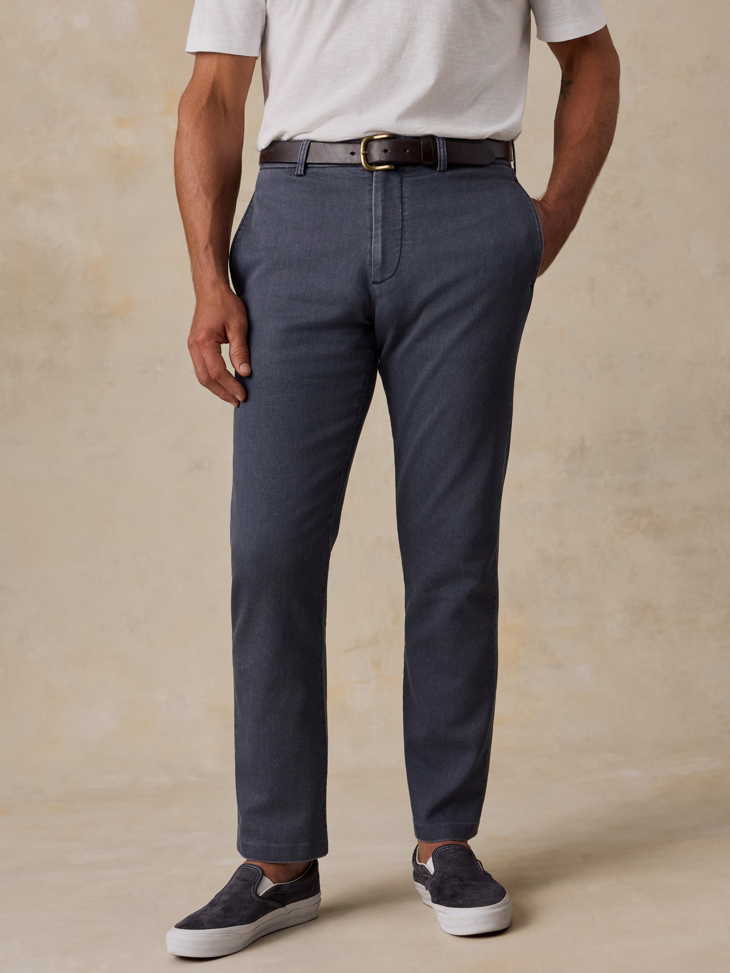 A man wearing slate blue slim-fit chino pants, styled with a white t-shirt, a dark brown leather belt, and navy slip-on sneakers, standing against a textured beige studio backdrop.