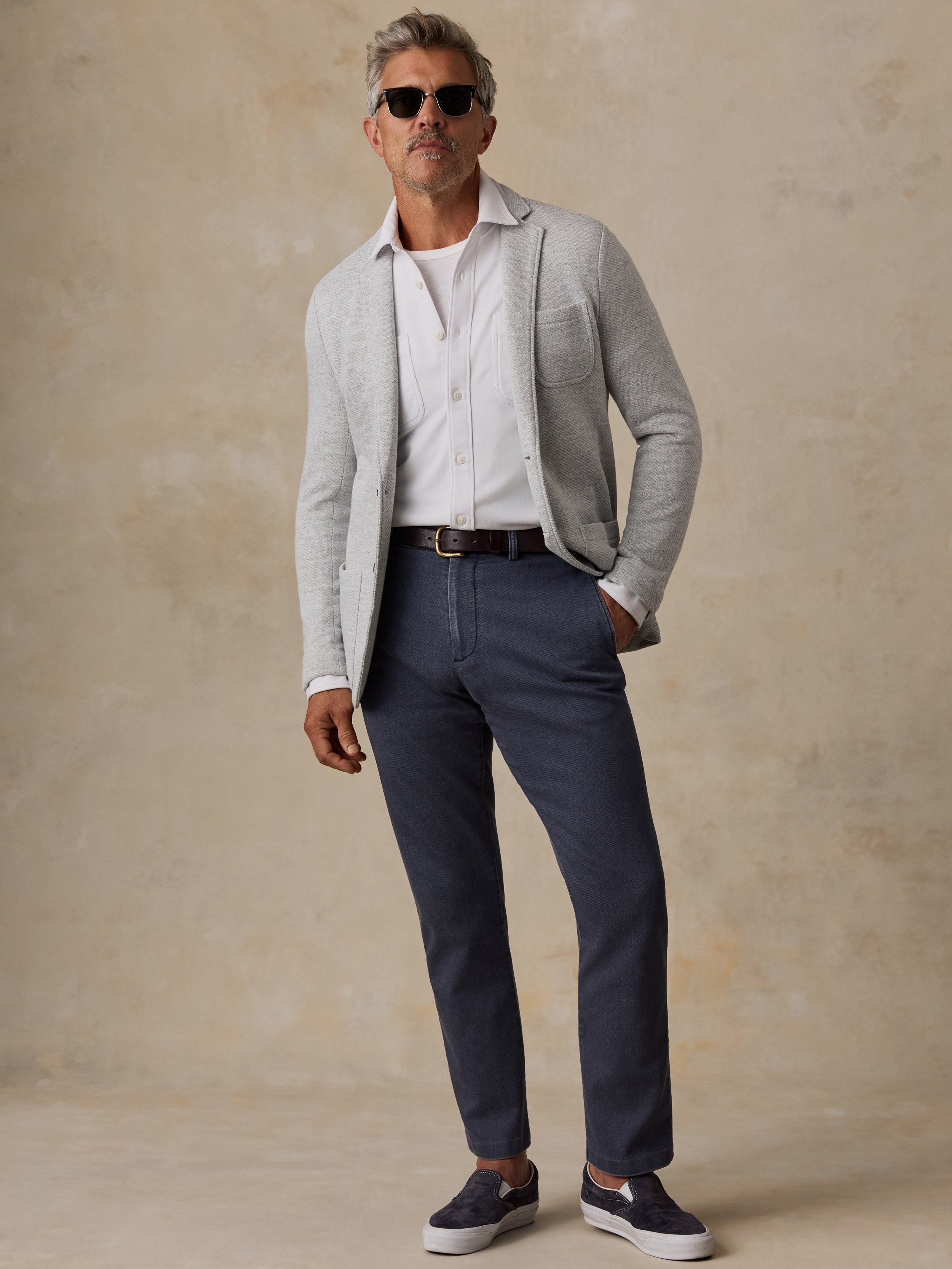 A man modeling a light gray knit blazer over a white button-down shirt, paired with navy blue trousers, a dark belt, and navy slip-on shoes, set against a textured beige studio backdrop.