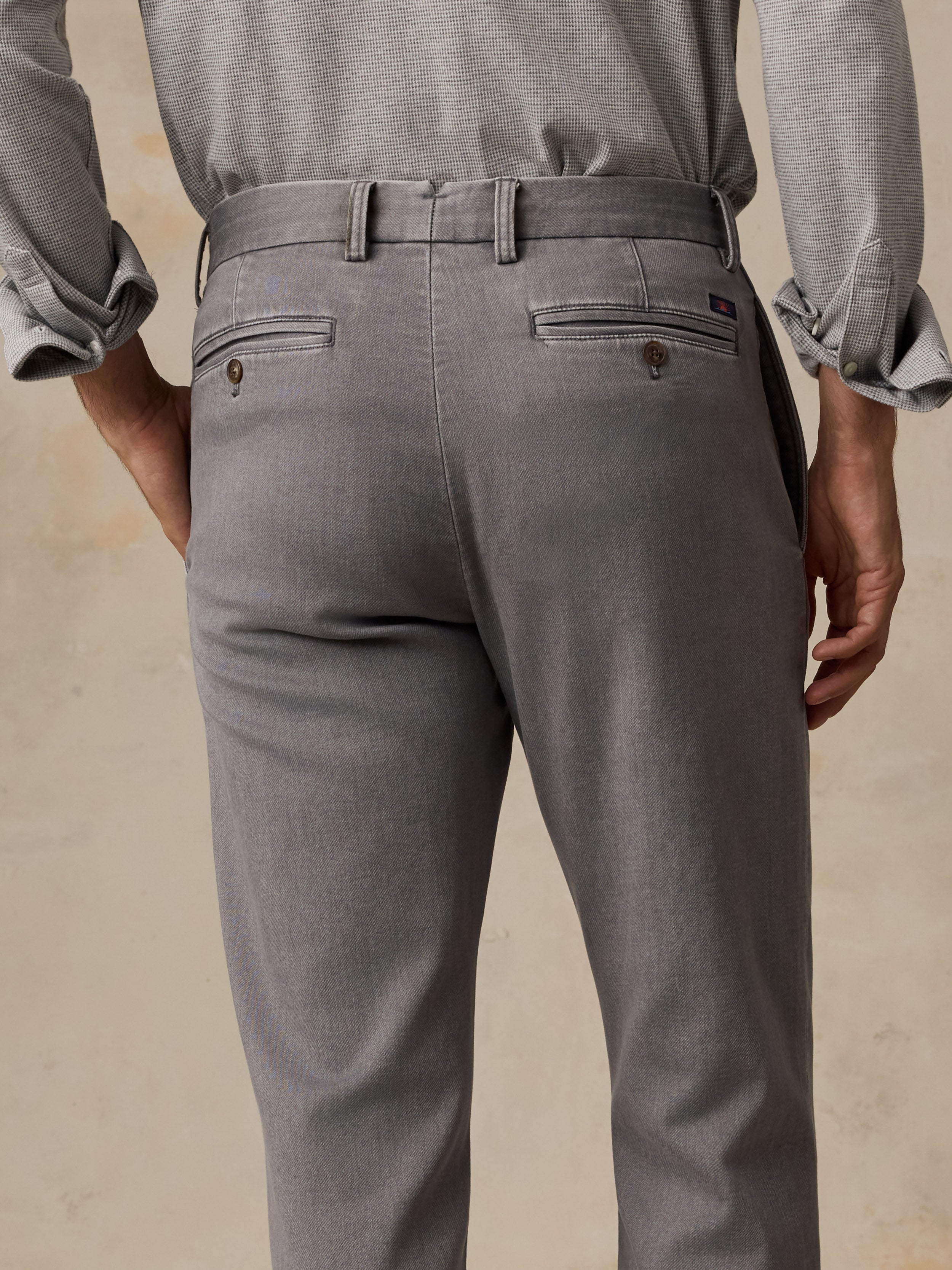 Rear view of a man wearing gray trousers with a matching belt, paired with a light gray long-sleeve shirt, against a neutral beige background.