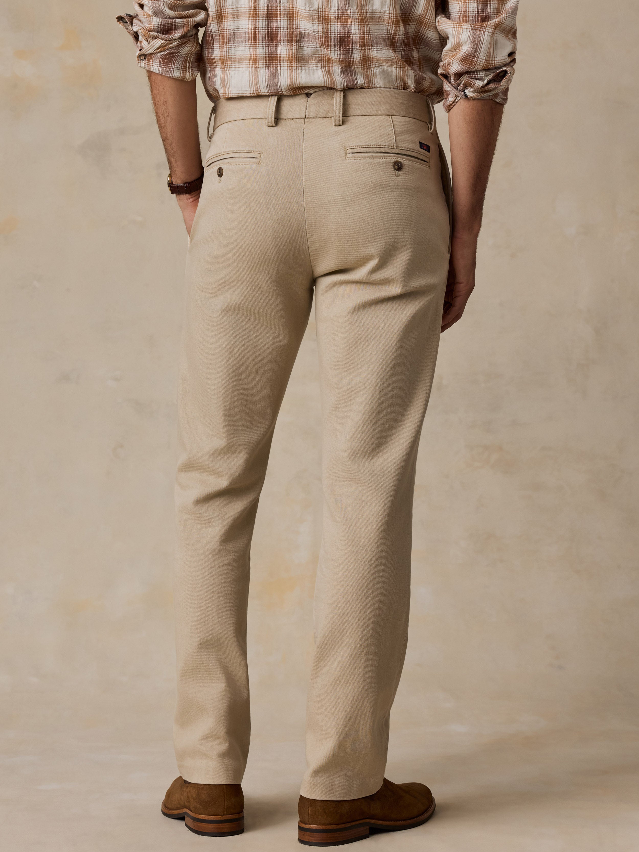 Rear view of a man wearing beige chino pants with a brown plaid shirt and brown shoes, standing against a textured beige studio backdrop.