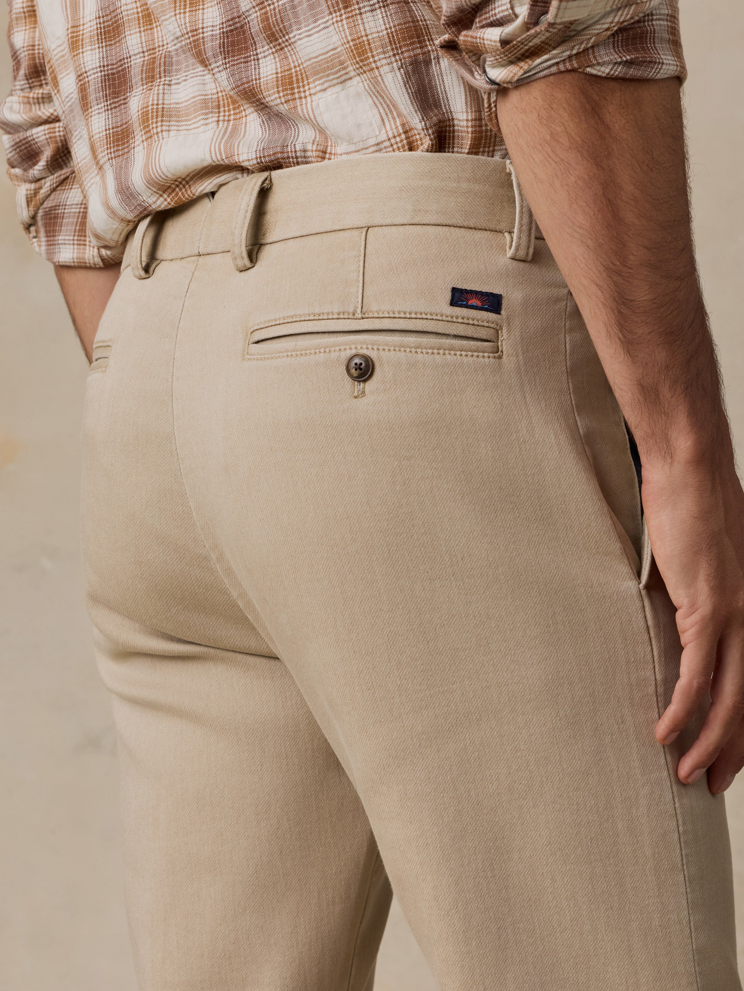 Close-up of a person wearing beige chino pants with a brown and beige plaid shirt tucked in, shown from the waist down against a light neutral background.