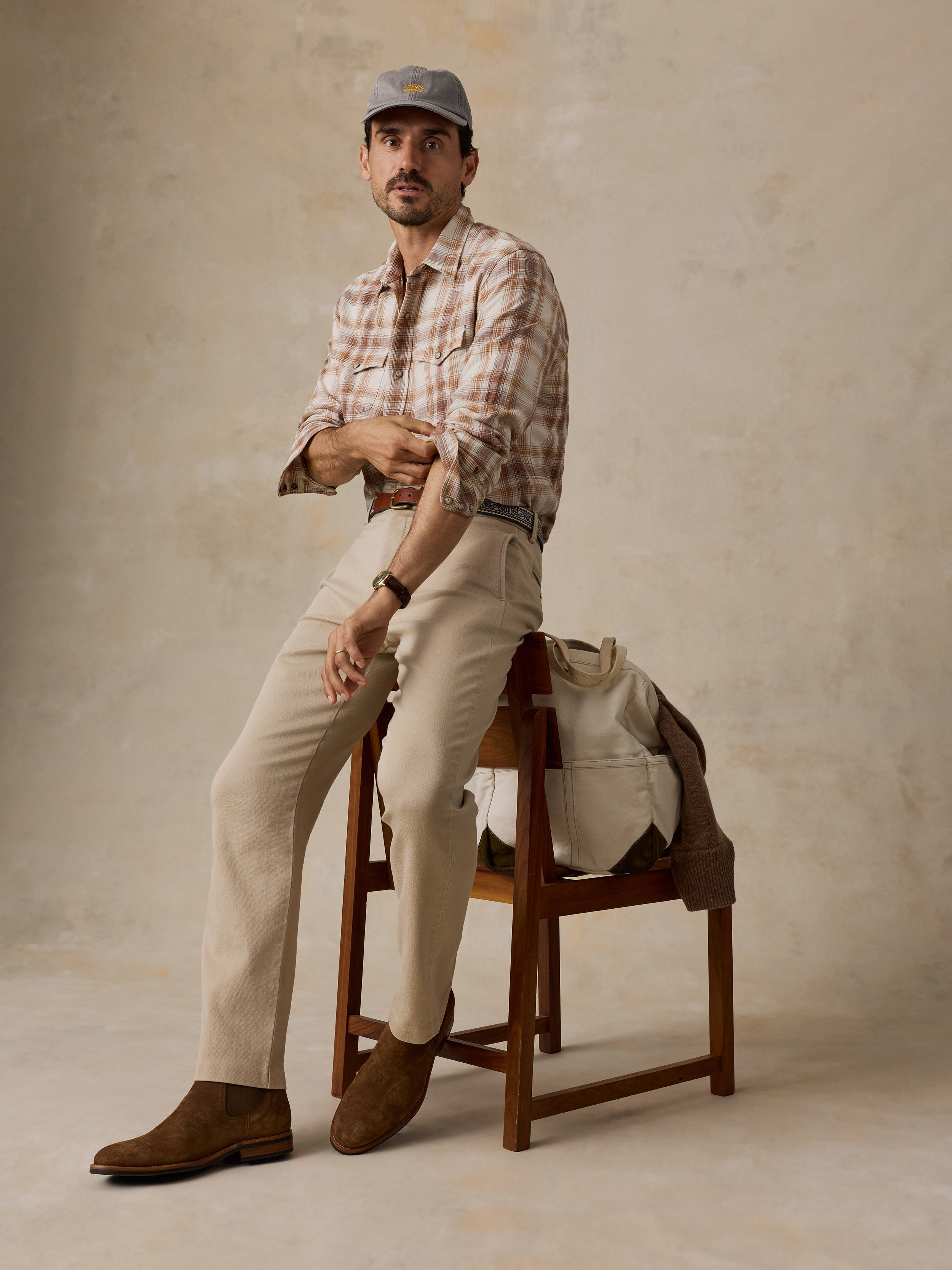 A man wearing a beige plaid shirt, beige trousers, brown suede boots, and a gray cap sits on a wooden stool against a textured beige studio backdrop.