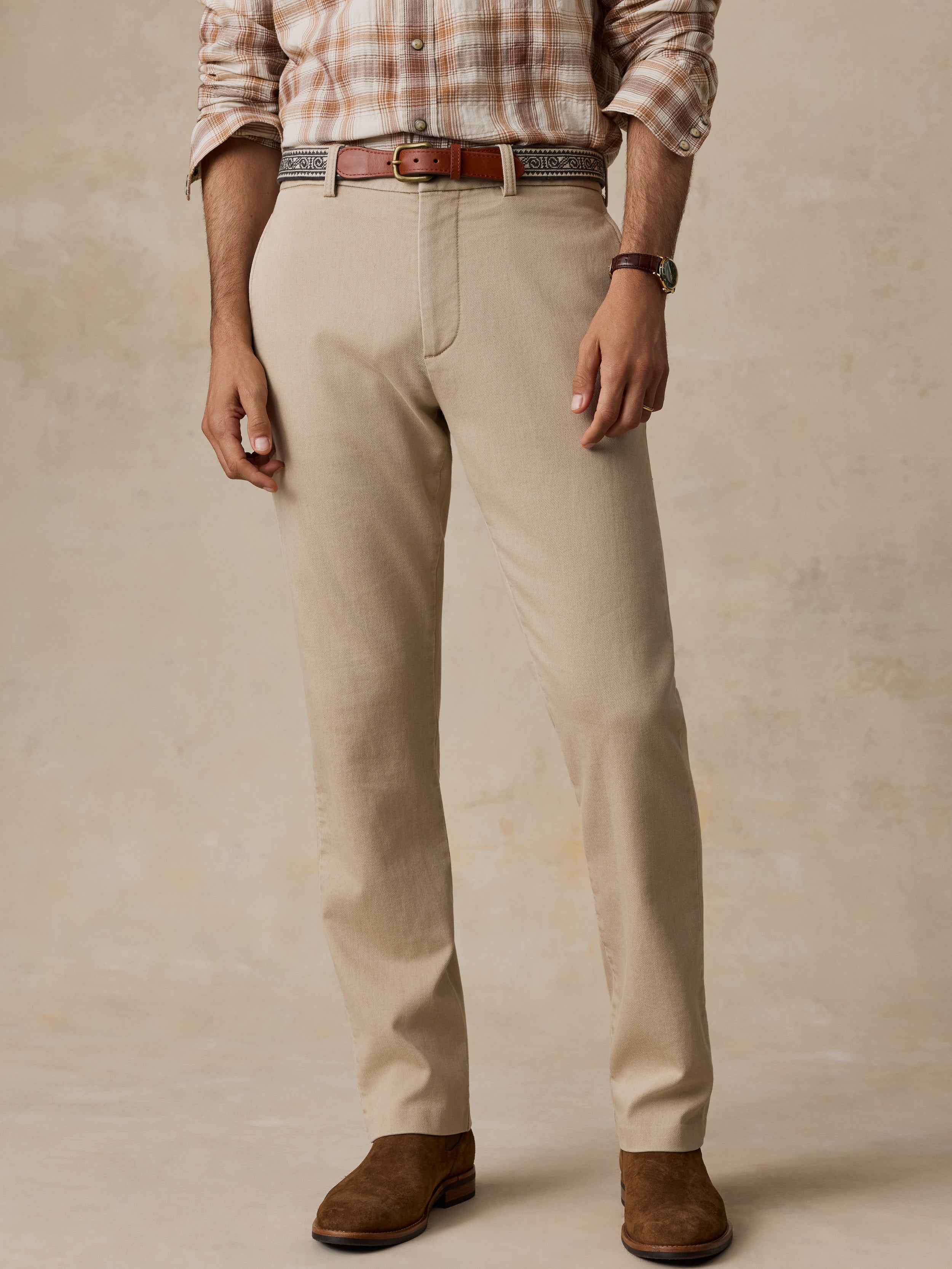 A man wearing beige chino pants, a brown leather belt, brown suede boots, and a brown and beige plaid shirt, standing against a textured beige studio backdrop.