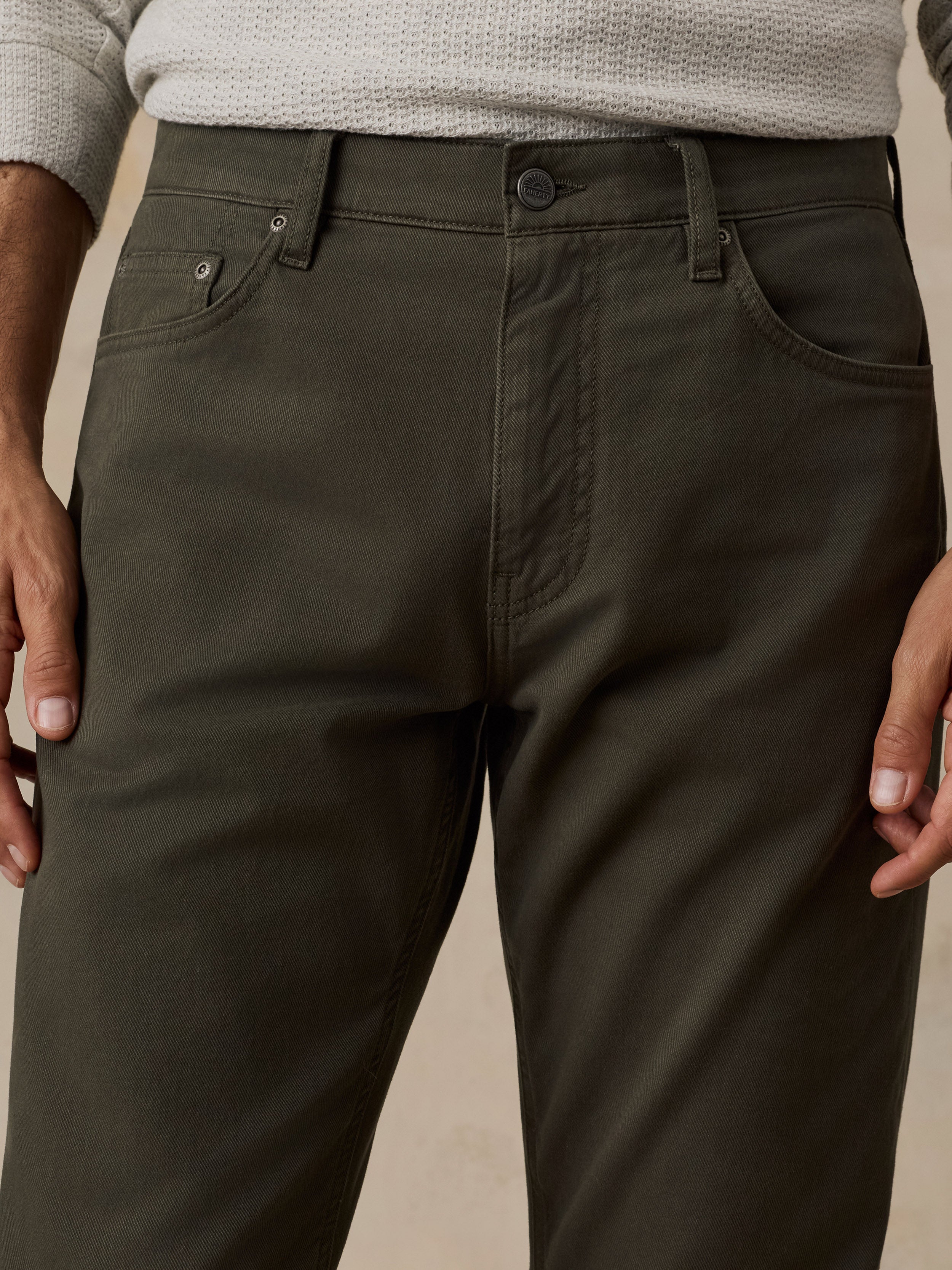 Close-up of a person wearing dark olive green trousers, showing the front waistband, belt loops, button, pockets, and hands against a plain beige background.
