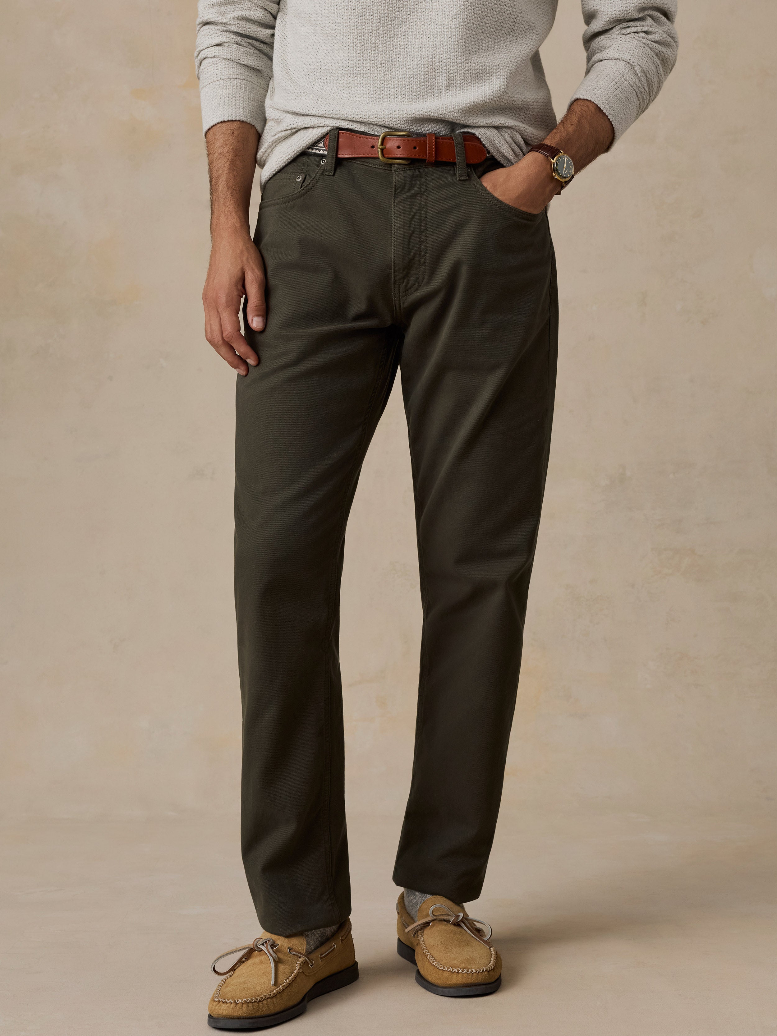 A man wearing dark olive green chinos with a brown leather belt, a light gray knit sweater, and tan suede boat shoes, standing against a textured beige background.