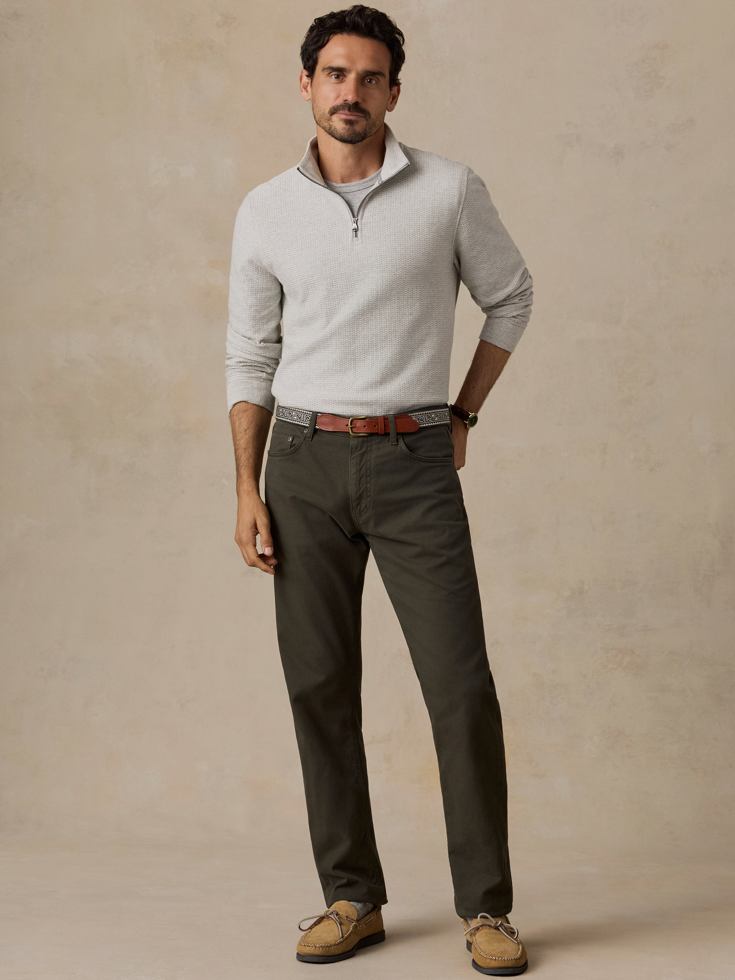 A man modeling a light gray quarter-zip knit sweater, dark olive green trousers, a brown leather belt, and tan boat shoes against a textured beige studio backdrop.
