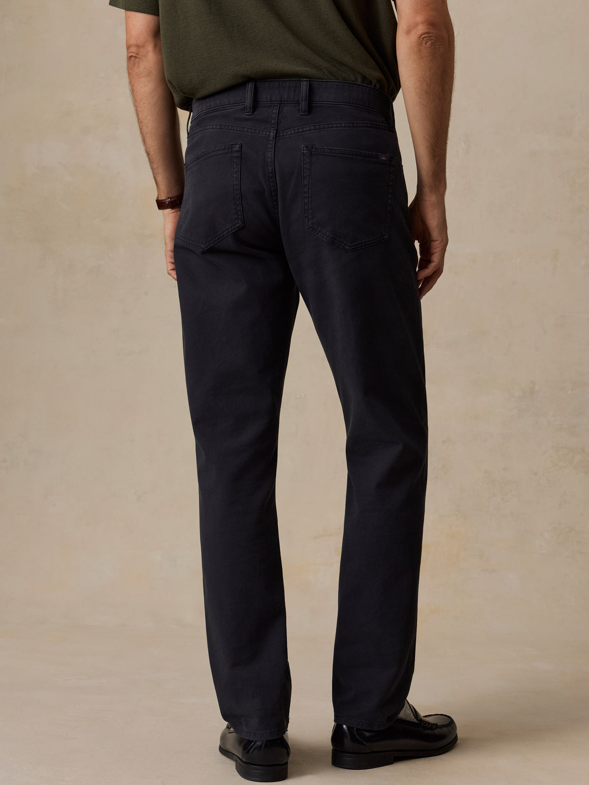 Rear view of a man wearing dark navy straight-leg trousers, a dark olive green t-shirt, and black leather shoes, standing against a textured beige studio backdrop., image 4 of 6