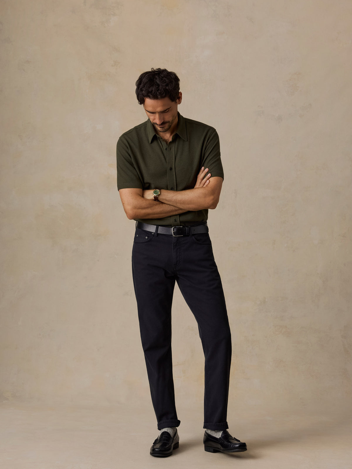 A man stands with arms crossed wearing a dark olive green short-sleeve button-up shirt, dark navy trousers, a black belt, and black loafers against a textured beige studio backdrop., image 3 of 6