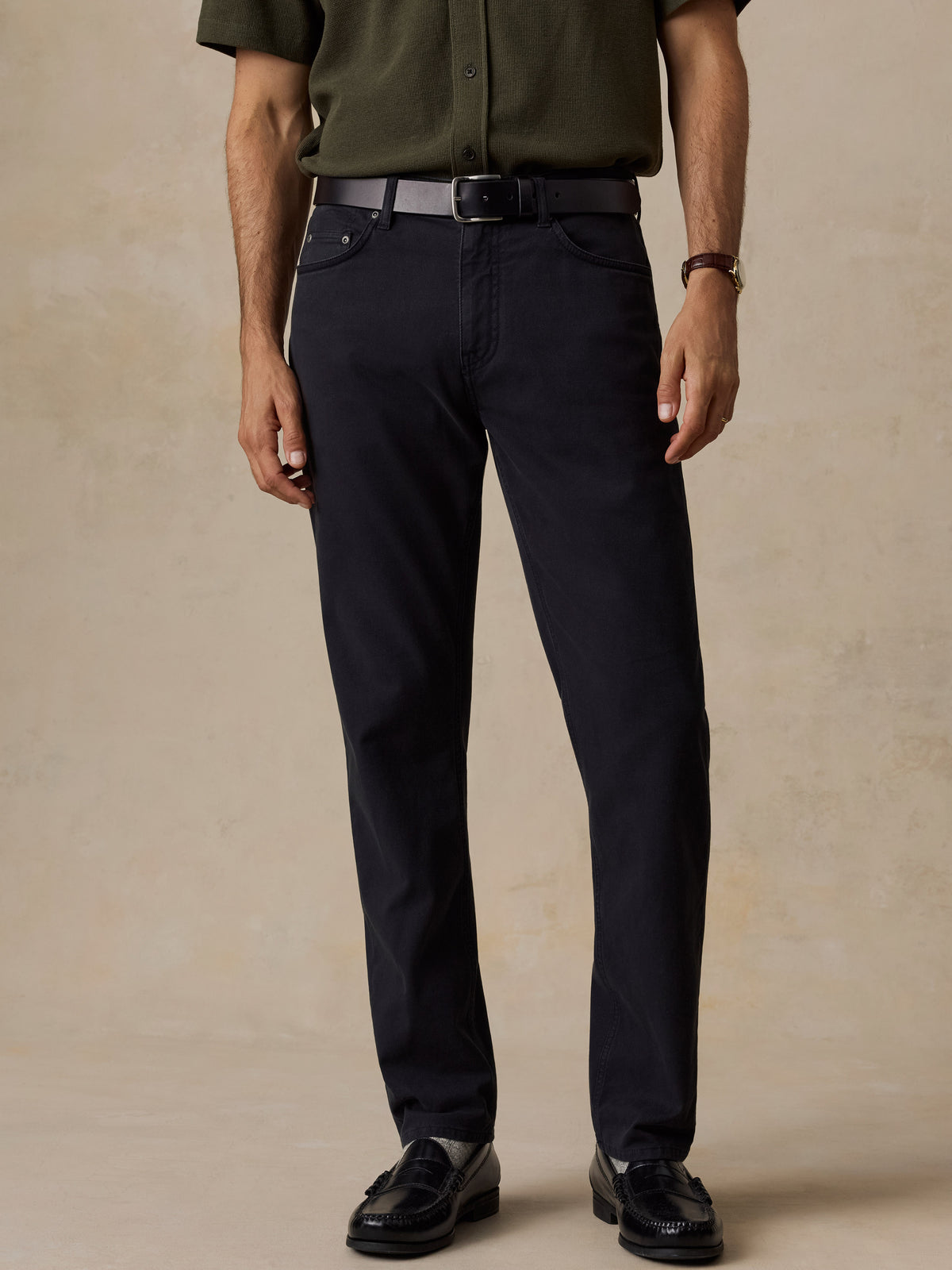 A man wearing dark navy blue slim-fit trousers, styled with an olive green shirt, a black belt, and black leather loafers, standing against a textured beige studio backdrop., image 1 of 6