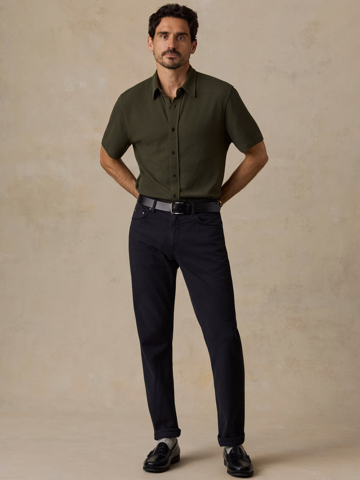 A man modeling a short-sleeve olive green button-down shirt and dark navy trousers, standing against a textured beige studio backdrop., image 6 of 6