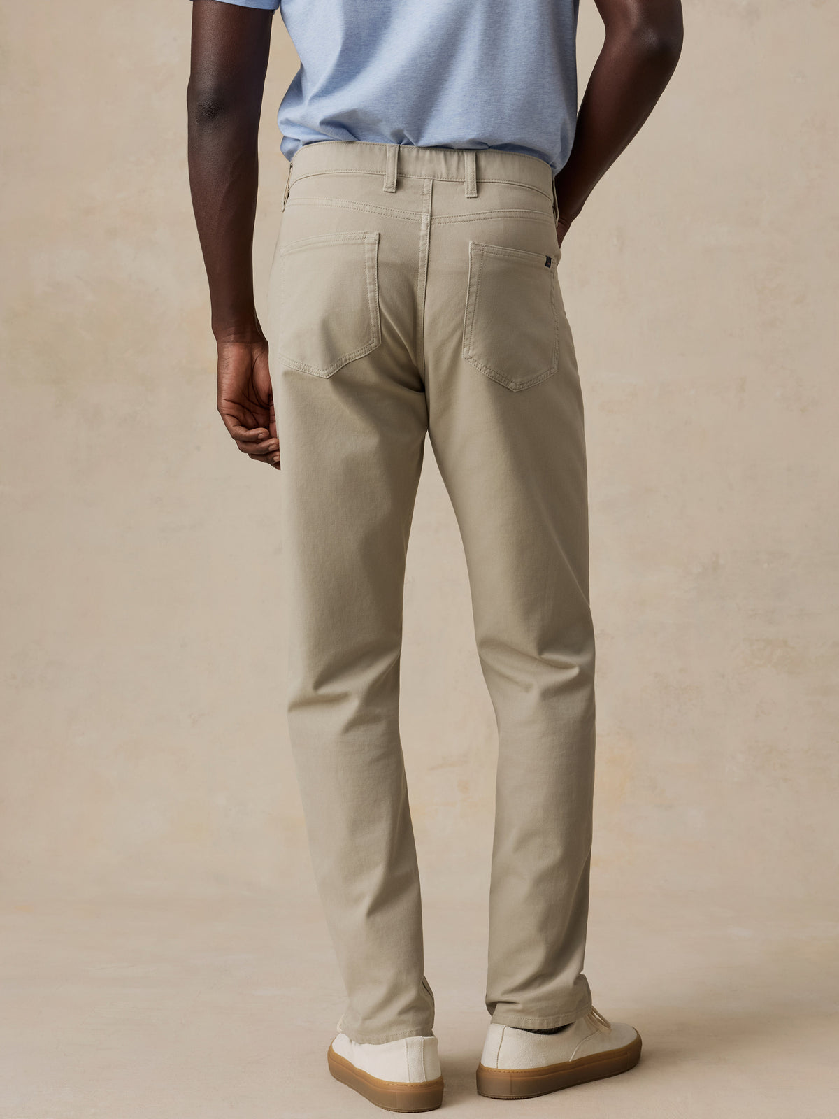A man shown from the back wearing light beige chino pants, a light blue t-shirt, and white slip-on shoes, standing against a textured beige studio backdrop., image 4 of 6