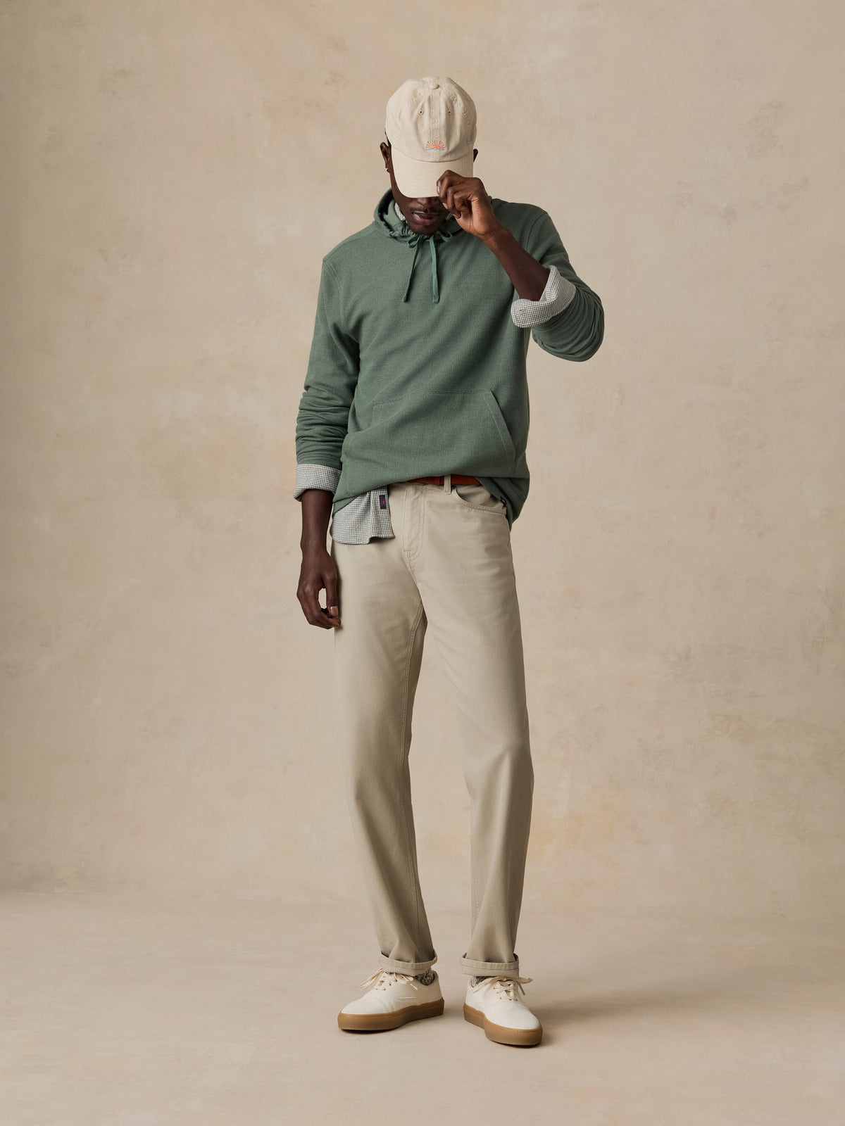 A model stands against a textured beige studio backdrop wearing a sage green long-sleeve polo shirt, light beige trousers, white sneakers, and a cream baseball cap., image 6 of 6