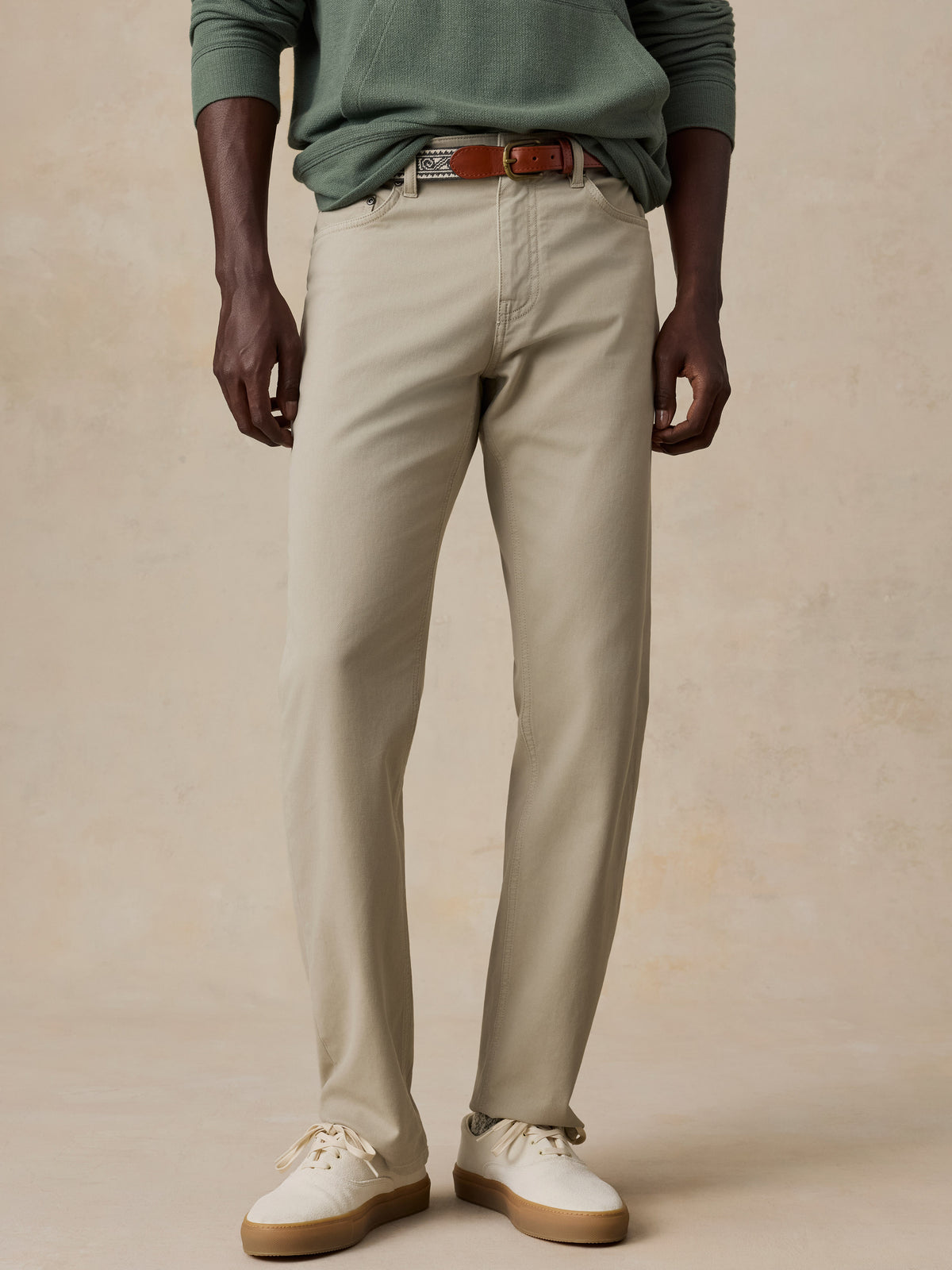 A model wearing a pair of light beige chinos with a brown leather belt, styled with a sage green long-sleeve shirt and off-white sneakers, set against a textured beige studio backdrop., image 1 of 6