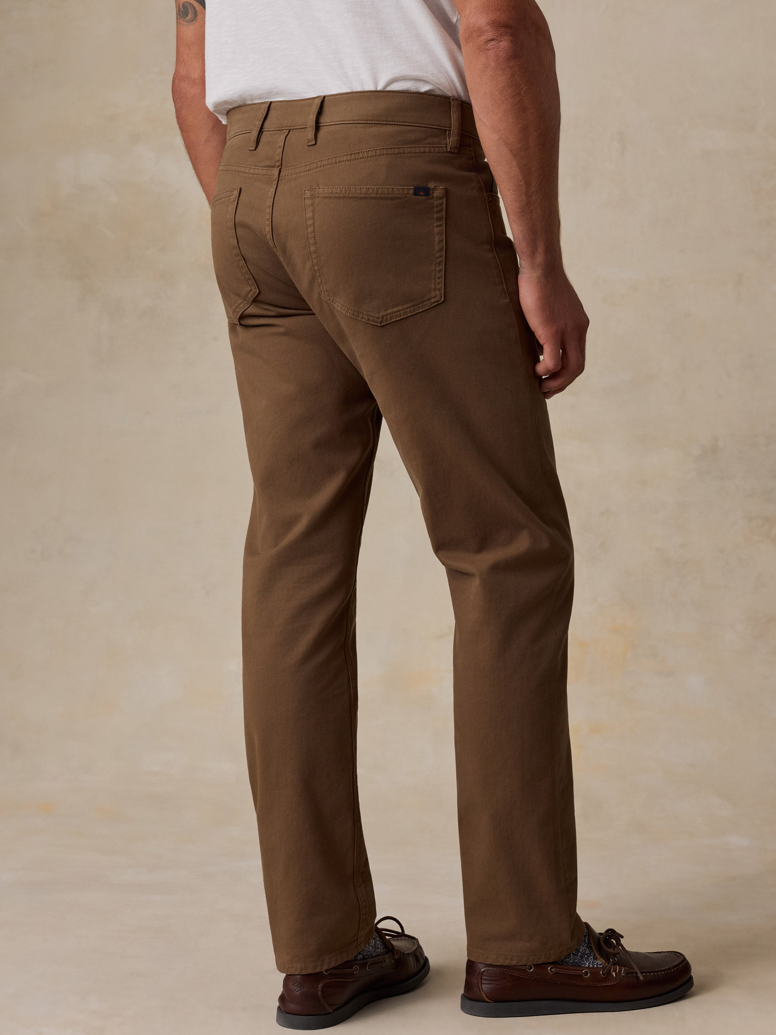 A man shown from the back wearing brown chino pants, a white t-shirt, and brown leather boat shoes, standing against a textured beige studio backdrop.