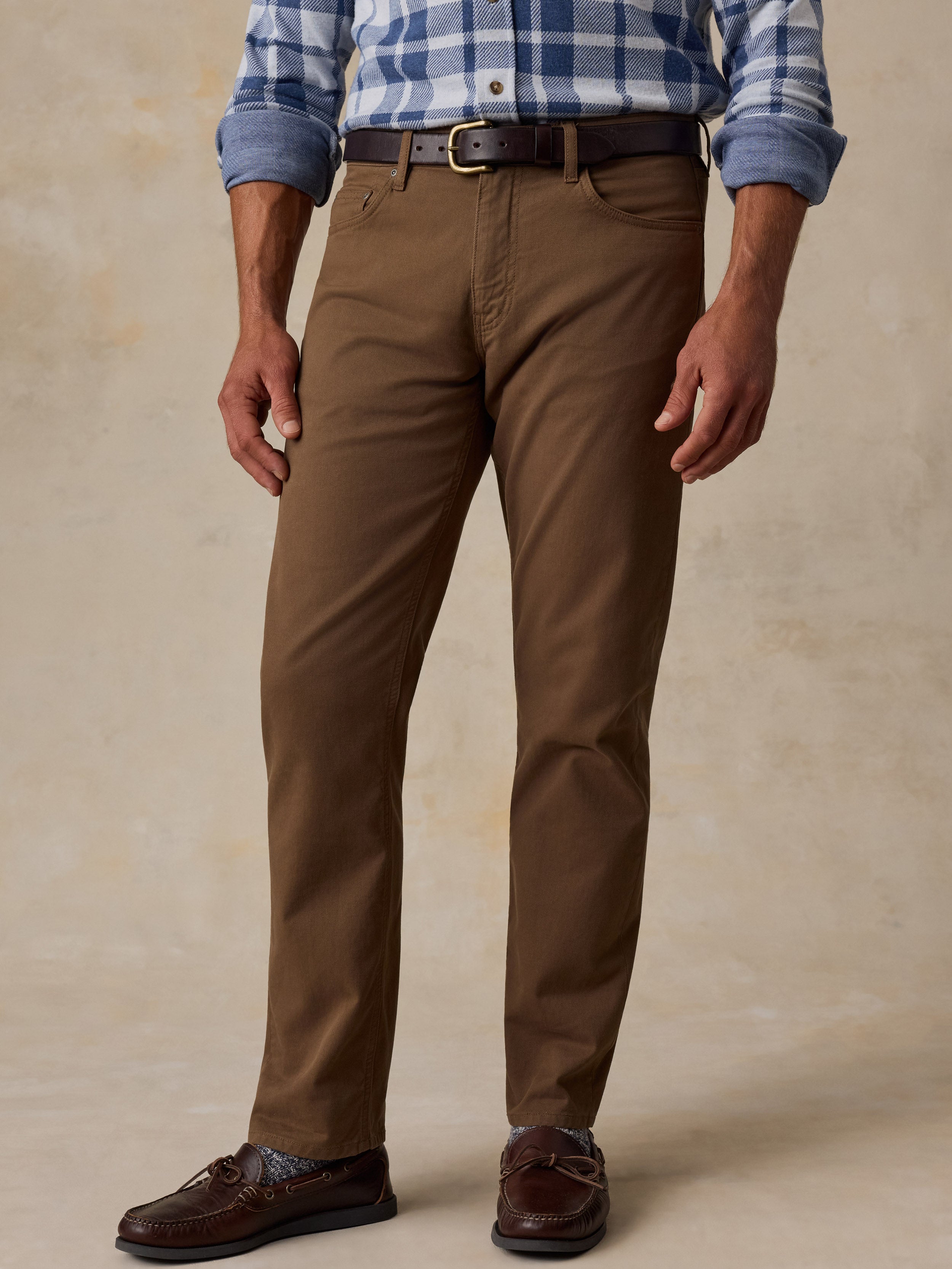 A man wearing brown chino pants, a blue and white plaid shirt, a dark brown belt, and brown leather loafers, standing against a textured beige studio backdrop.