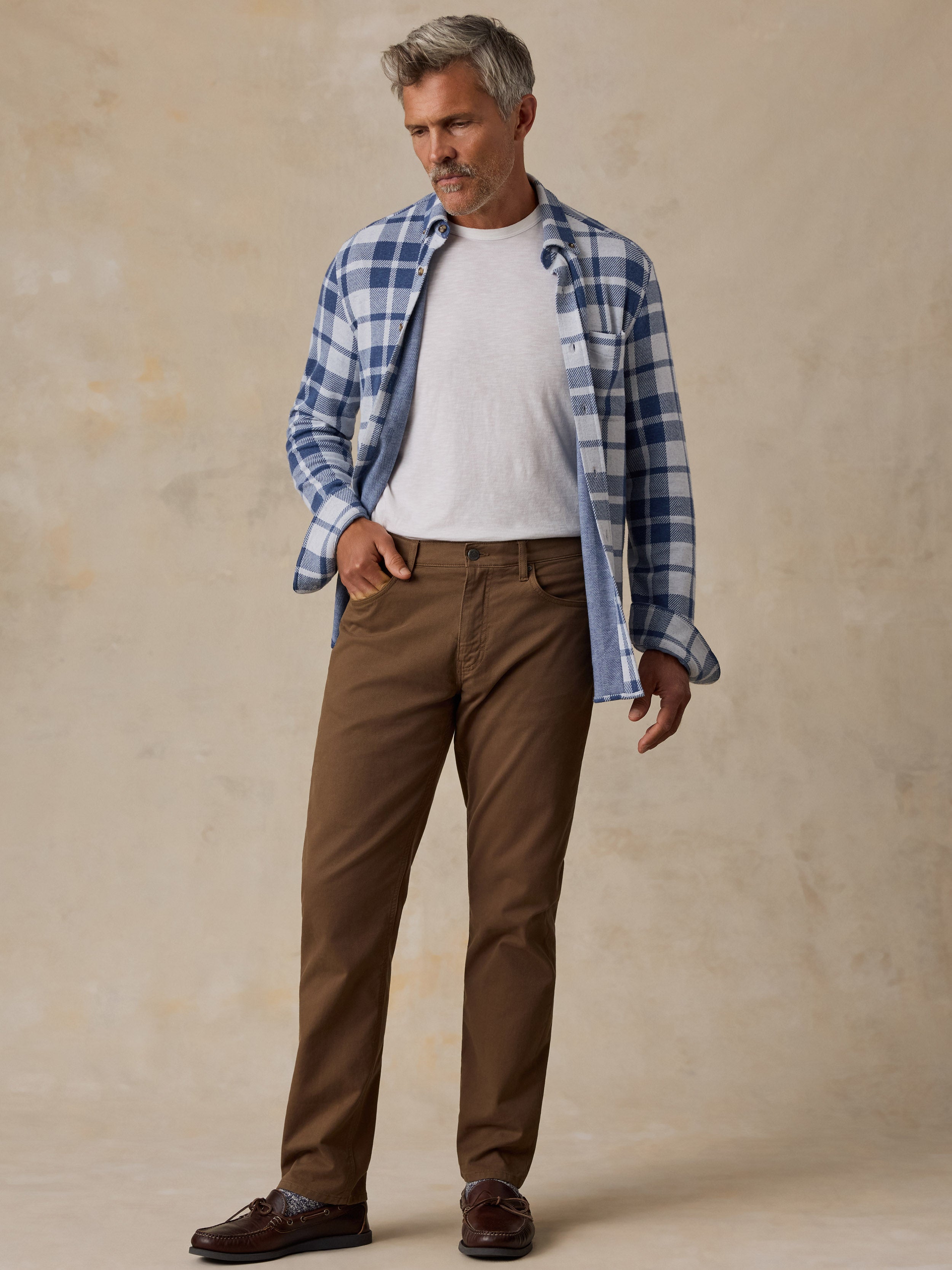 A man modeling a blue and white plaid button-down shirt over a white t-shirt, paired with brown trousers and dark brown leather shoes, standing against a textured beige studio backdrop.