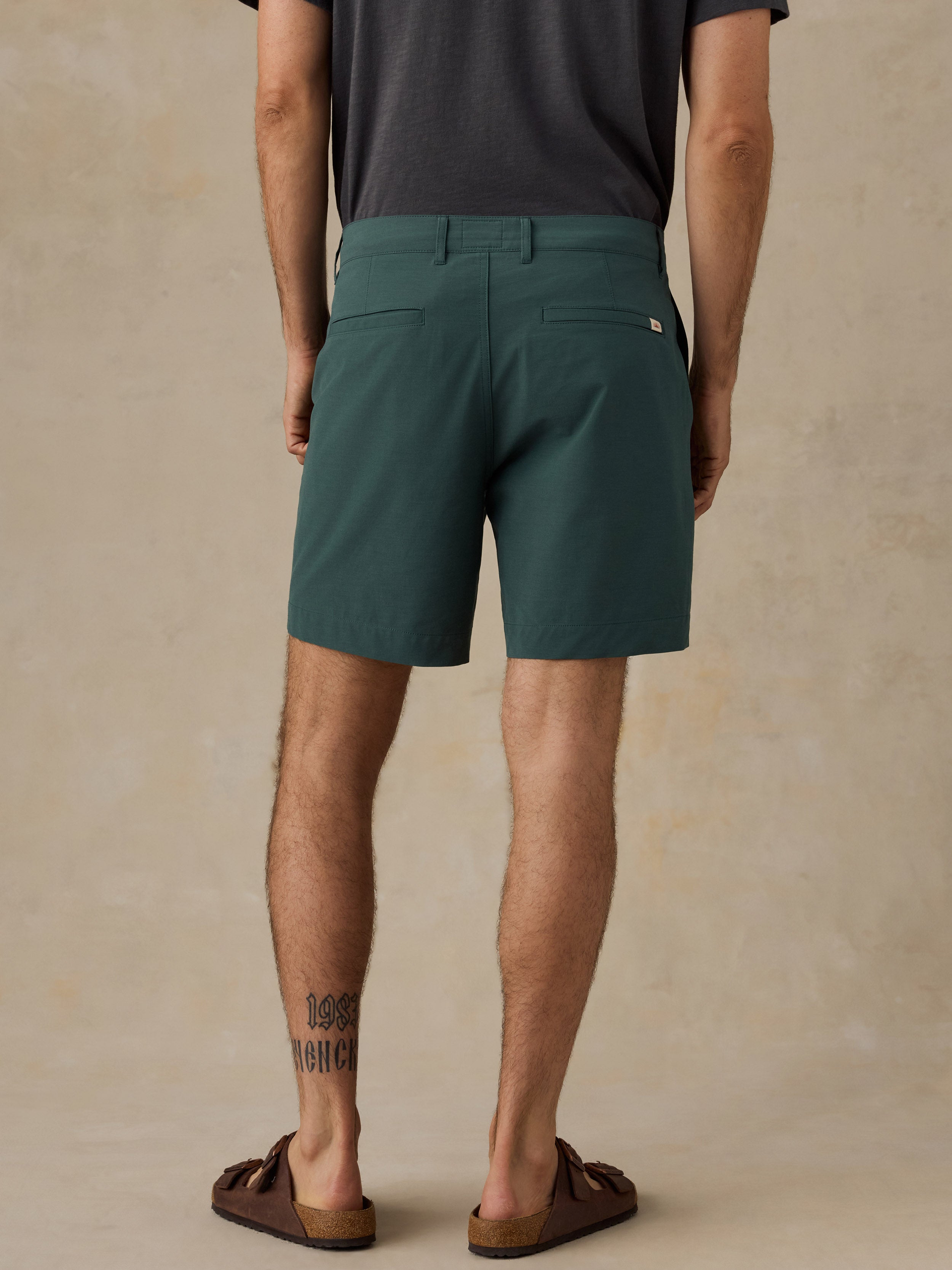 A man viewed from the back wearing dark teal-green shorts, a dark gray t-shirt, and brown slide sandals, standing against a textured beige studio backdrop.